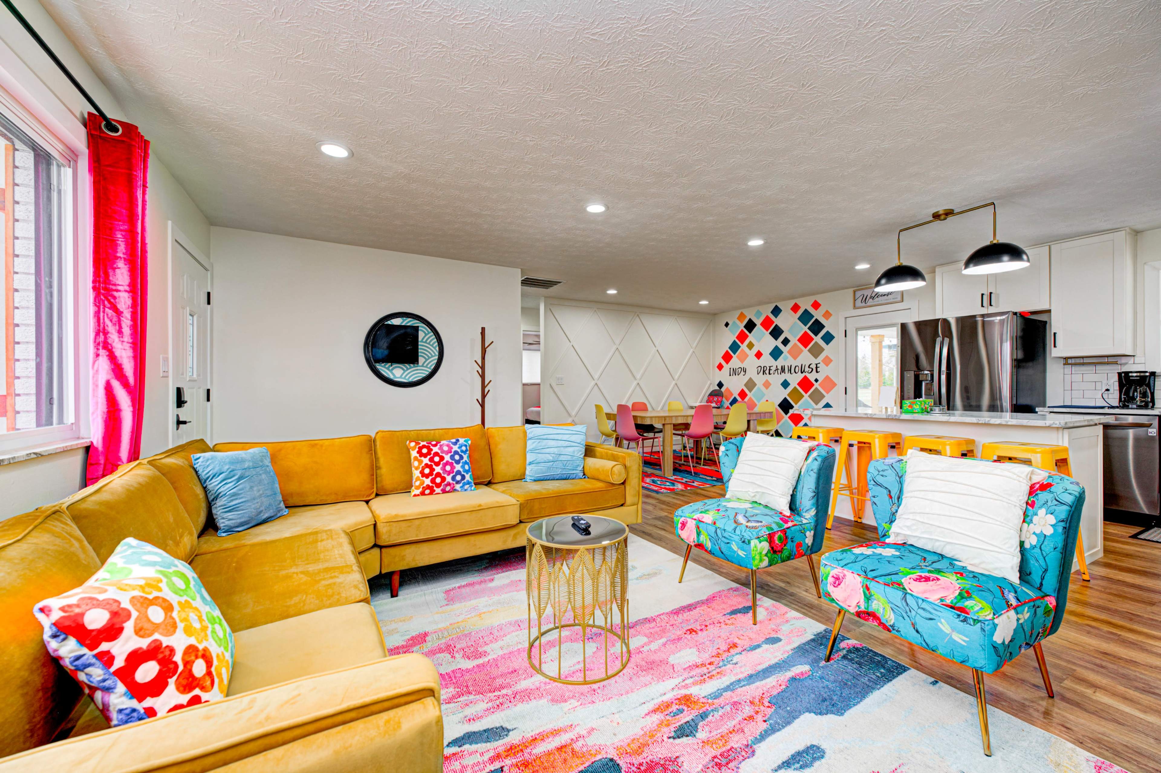 The image shows a colorful living area with a yellow sectional sofa, patterned chairs, and a vibrant rug, adjoining a dining space with a mural.