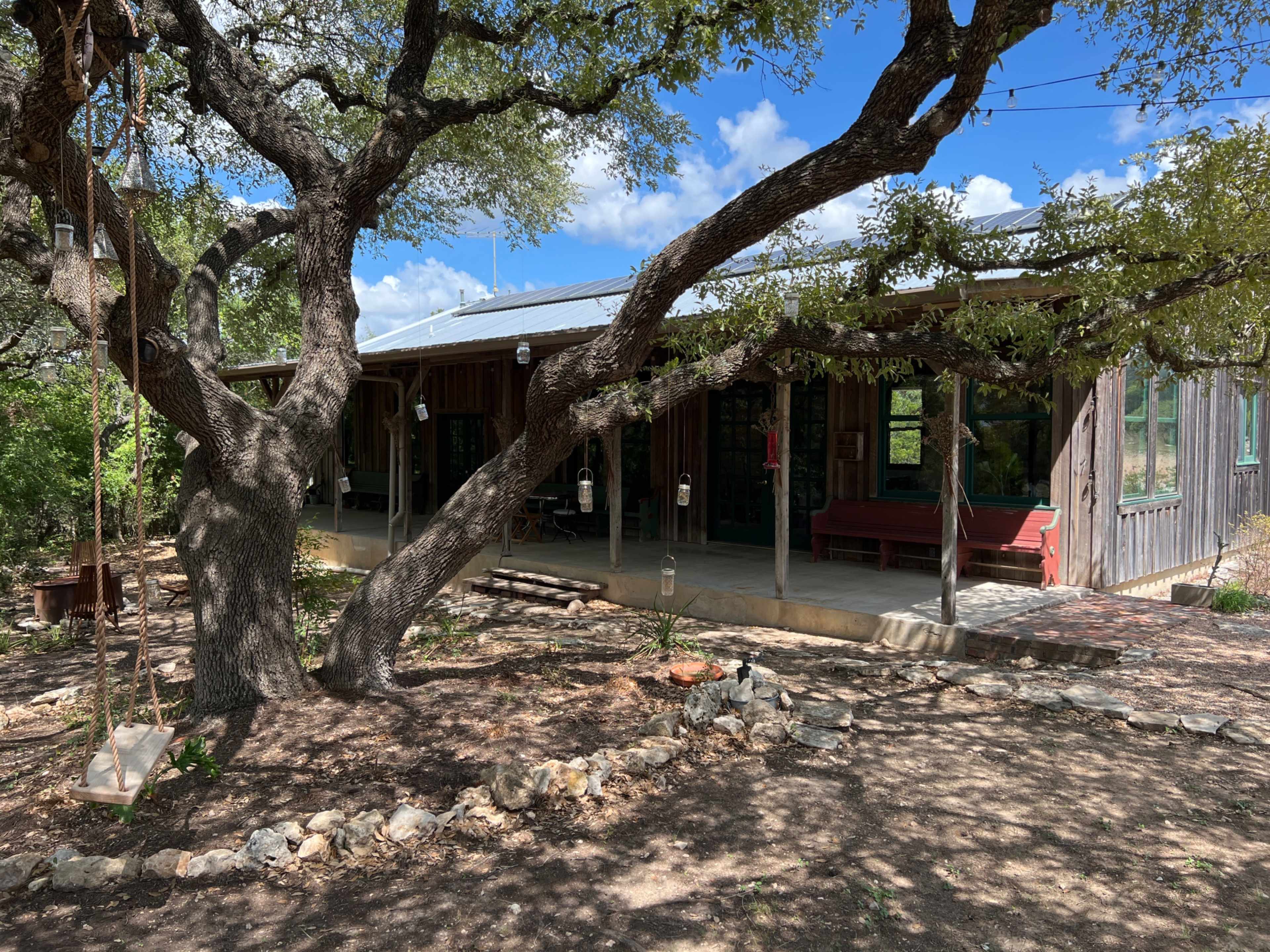 A rustic wooden house stands under the shade of large trees, with a stone pathway leading to its entrance.