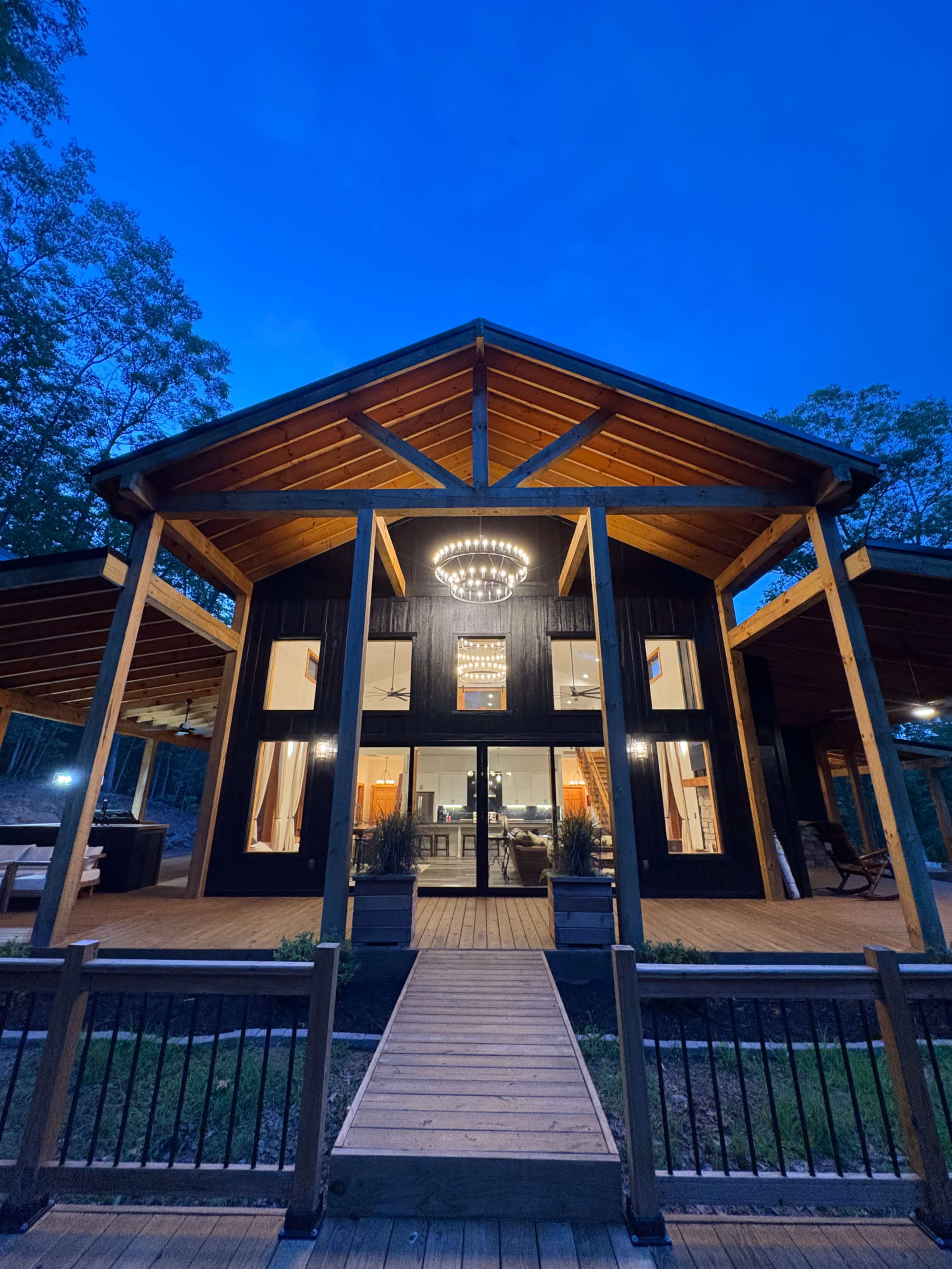A modern, dark-colored house with a peaked roof and large front windows is illuminated by a chandelier, surrounded by trees at dusk.