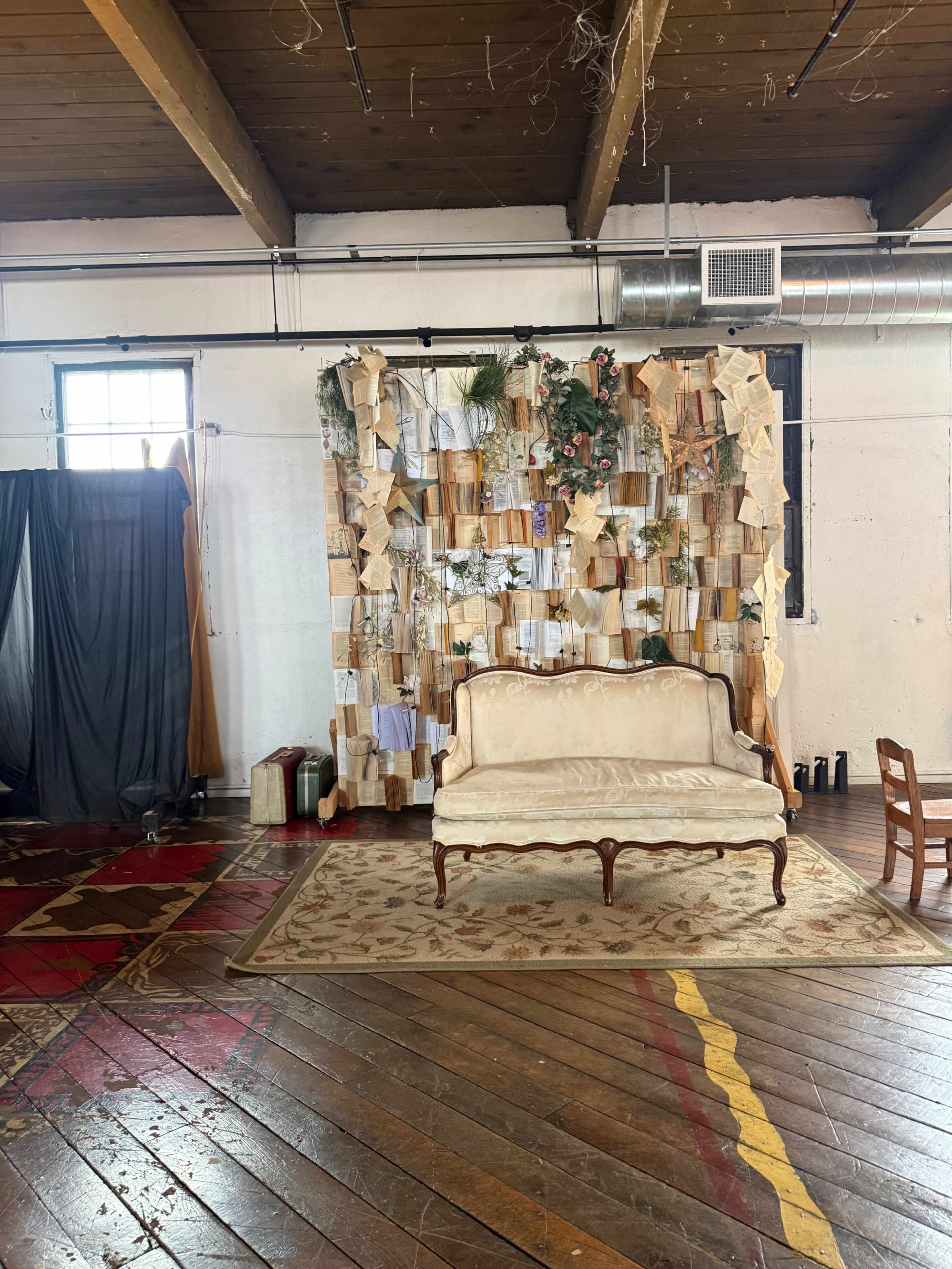 The image shows a vintage sofa in front of a textured wall adorned with fabric swatches and greenery within a spacious room featuring wooden floors.