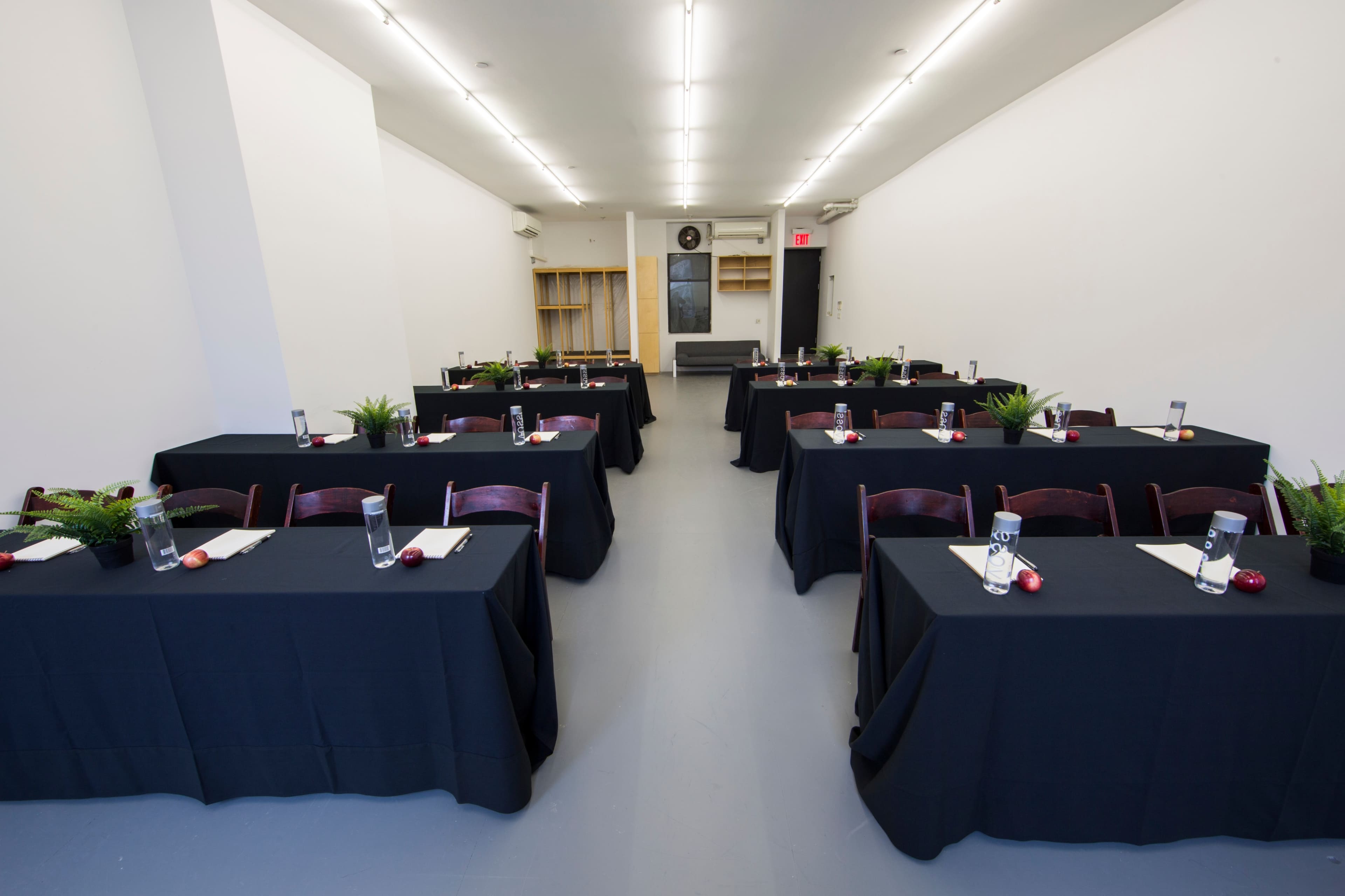 A spacious conference room features multiple tables covered with black linens, each set with water glasses and small plants.