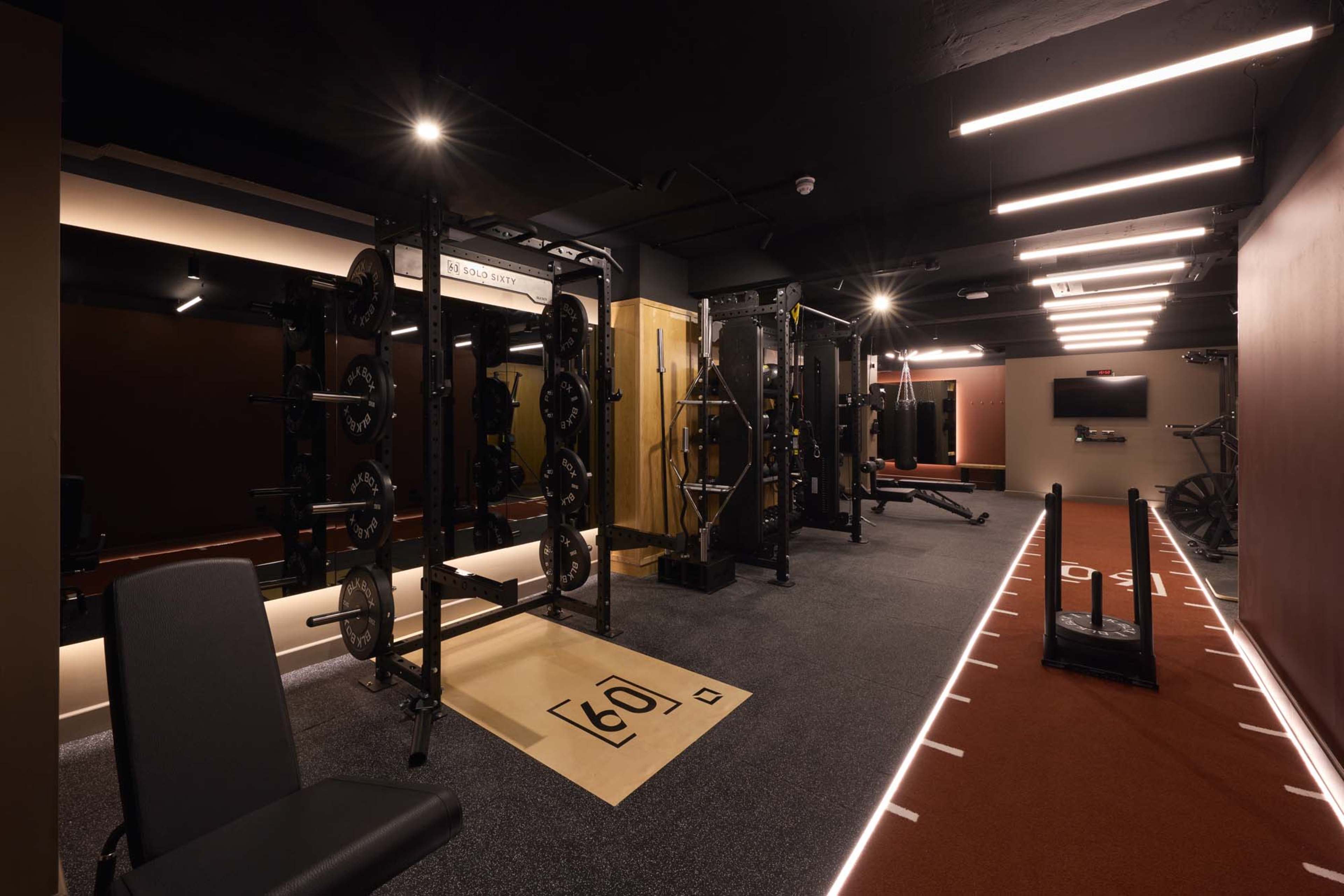 The image shows a modern gym interior featuring weightlifting equipment, a cardio area with a treadmill, and a training track marked with lane lines.