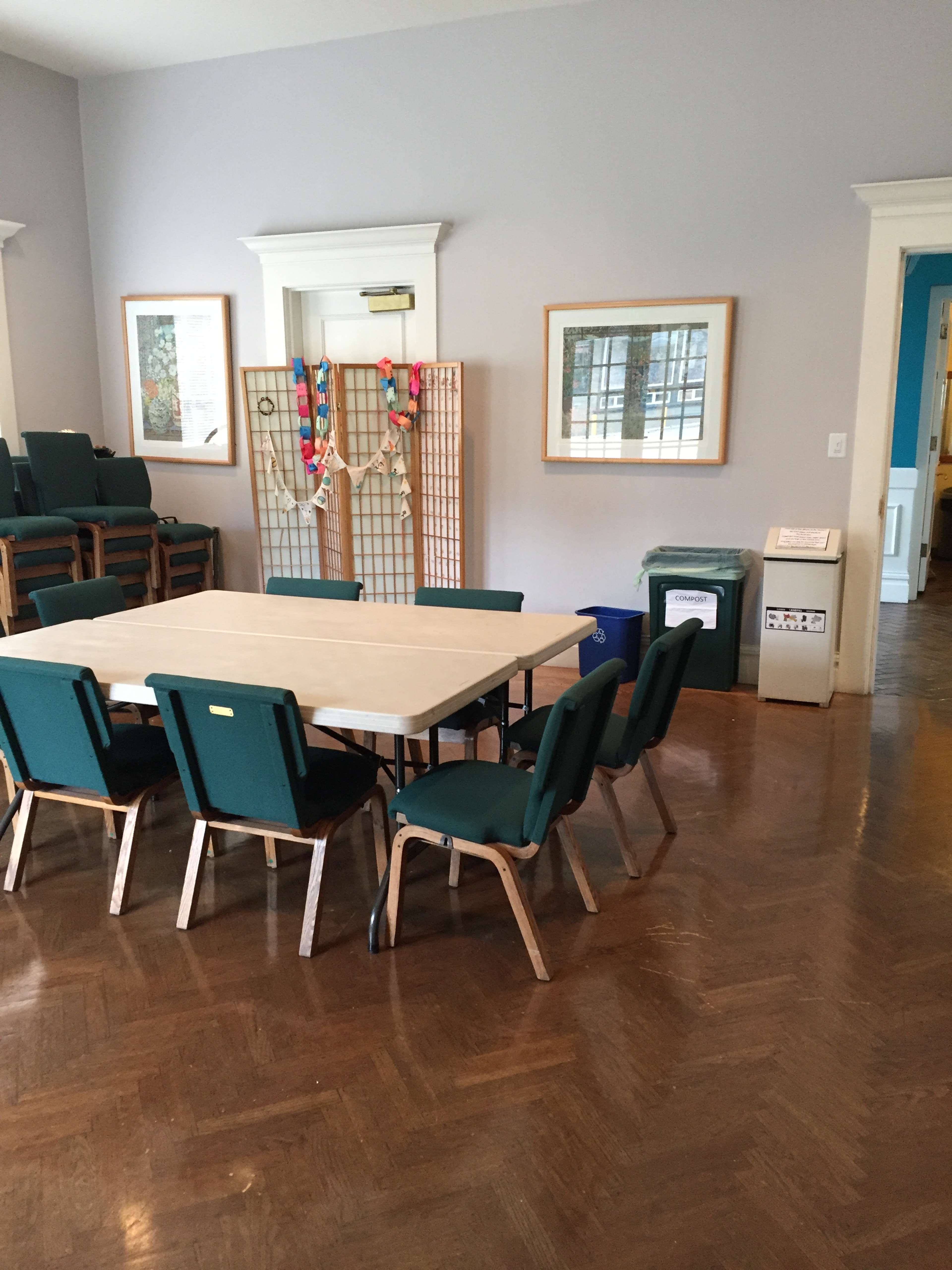 A square table surrounded by eight green chairs is positioned in a room with light-colored walls and wooden flooring.