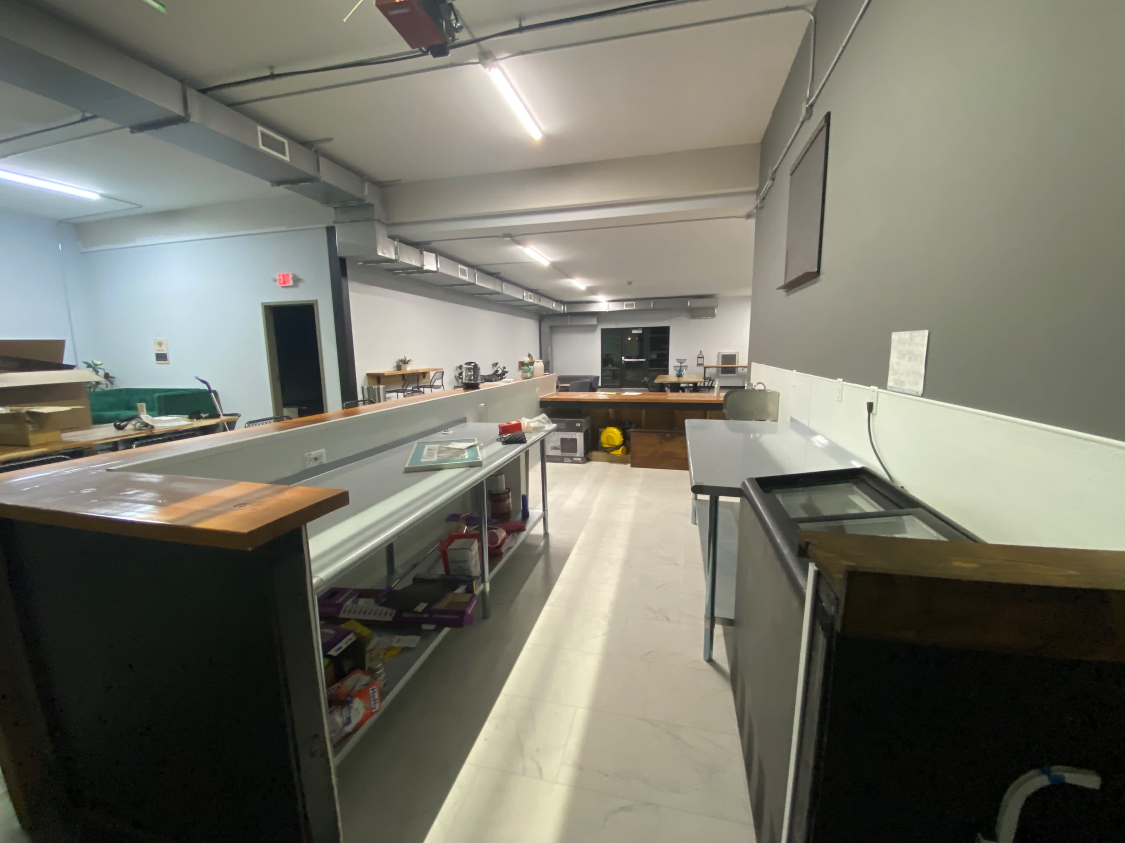 The image shows a large, modern kitchen space with stainless steel countertops, white walls, and various appliances set up along the perimeter.