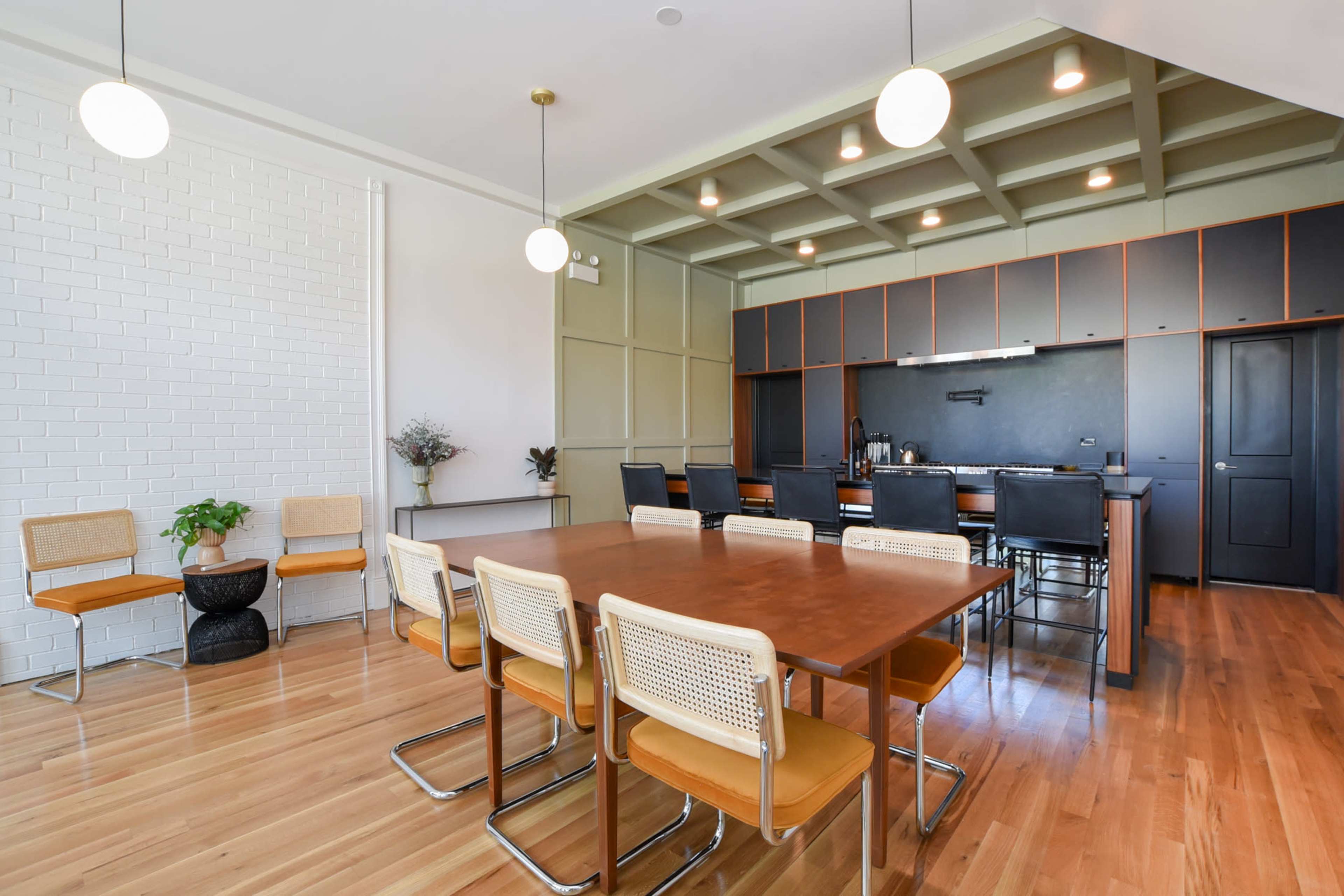 A modern dining area with a wooden table surrounded by metal-framed chairs, adjacent to a kitchen featuring dark cabinetry and overhead lighting.