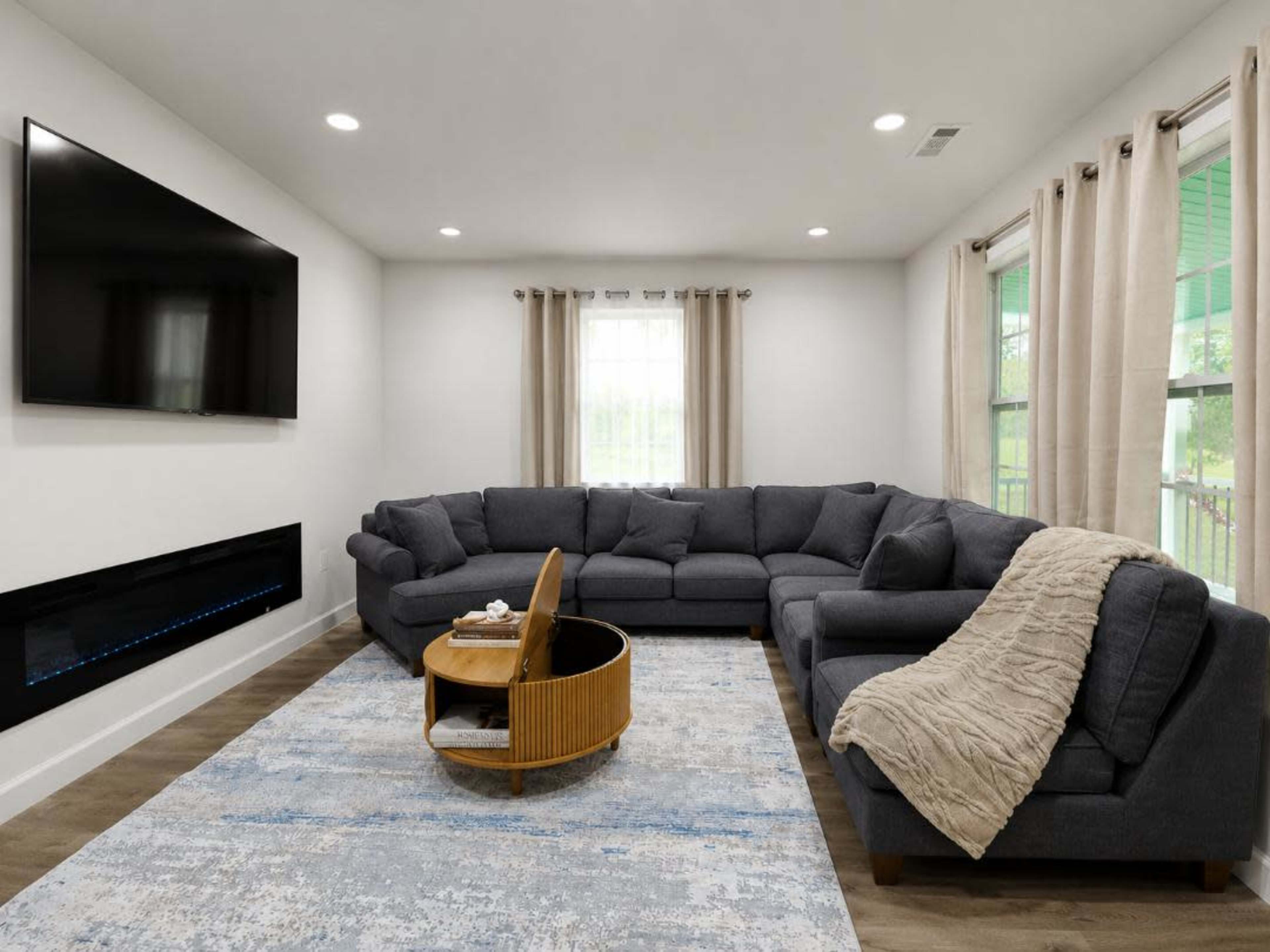 The image depicts a modern living room featuring a large sectional sofa, a circular coffee table, a wall-mounted television, and a sleek electric fireplace, with natural light coming through large windows.
