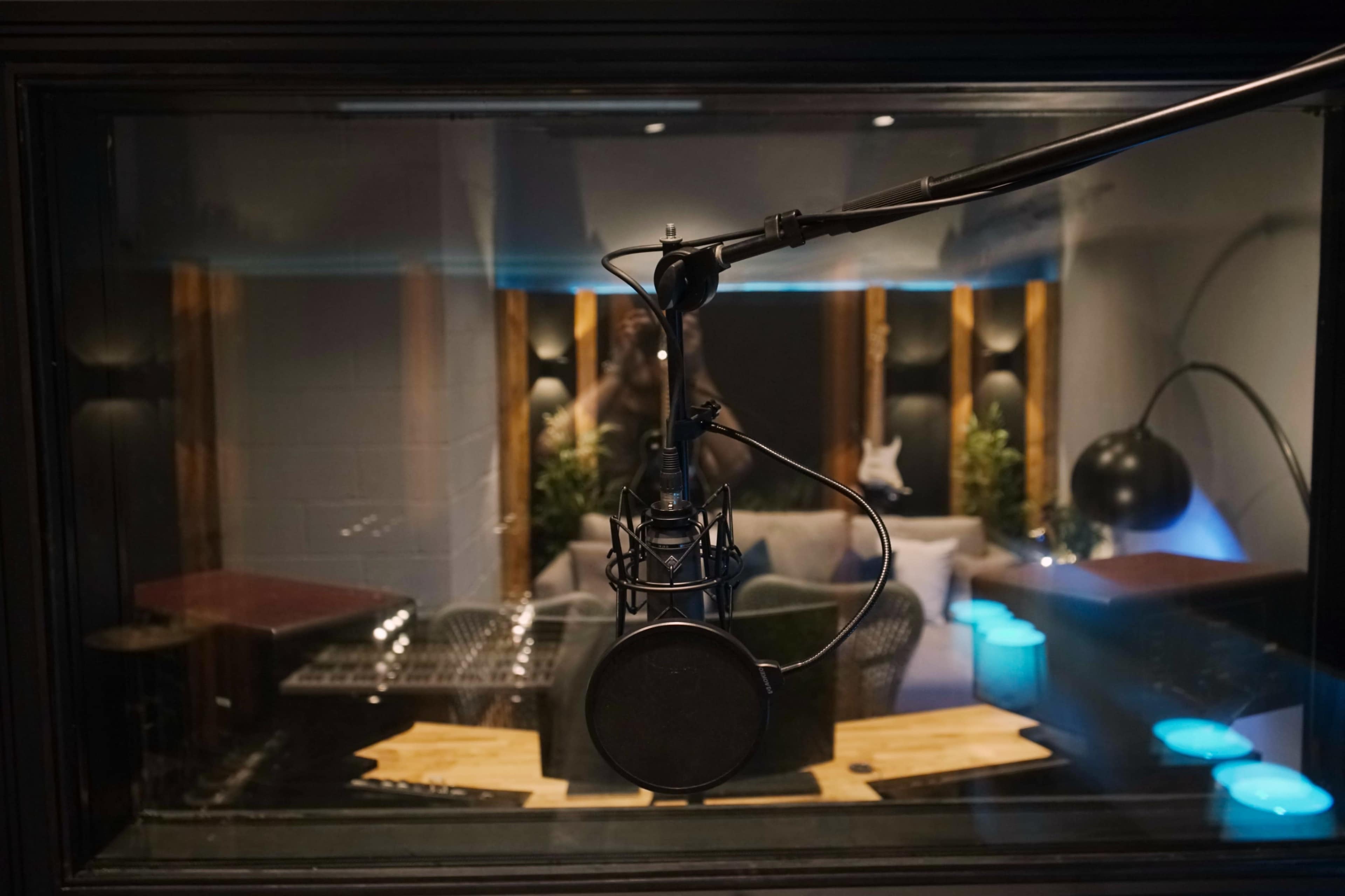 The image shows a microphone suspended in a recording studio, with a cozy seating area visible in the background.