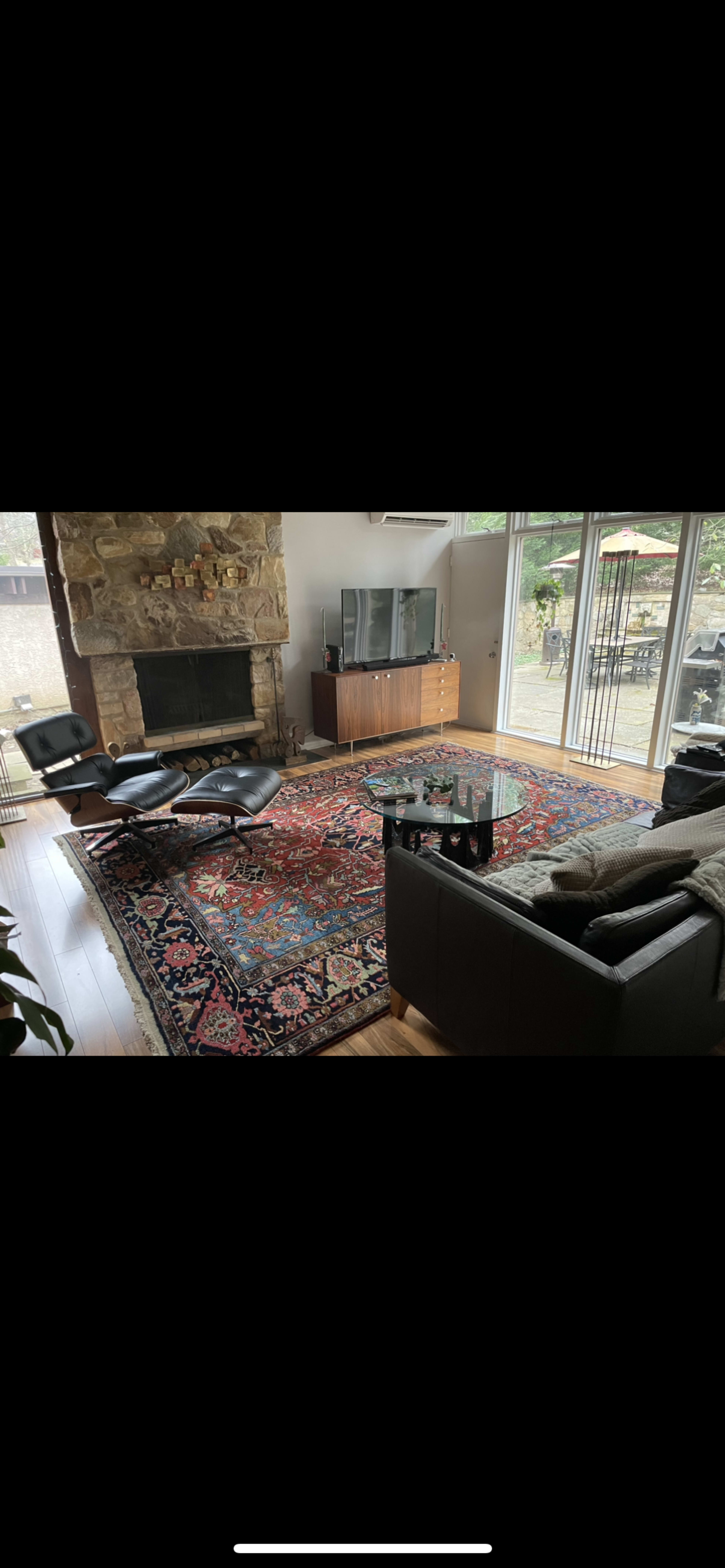 A living room features a stone fireplace, a large area rug, a black sofa, three lounge chairs, a glass coffee table, and a television on a wooden console.