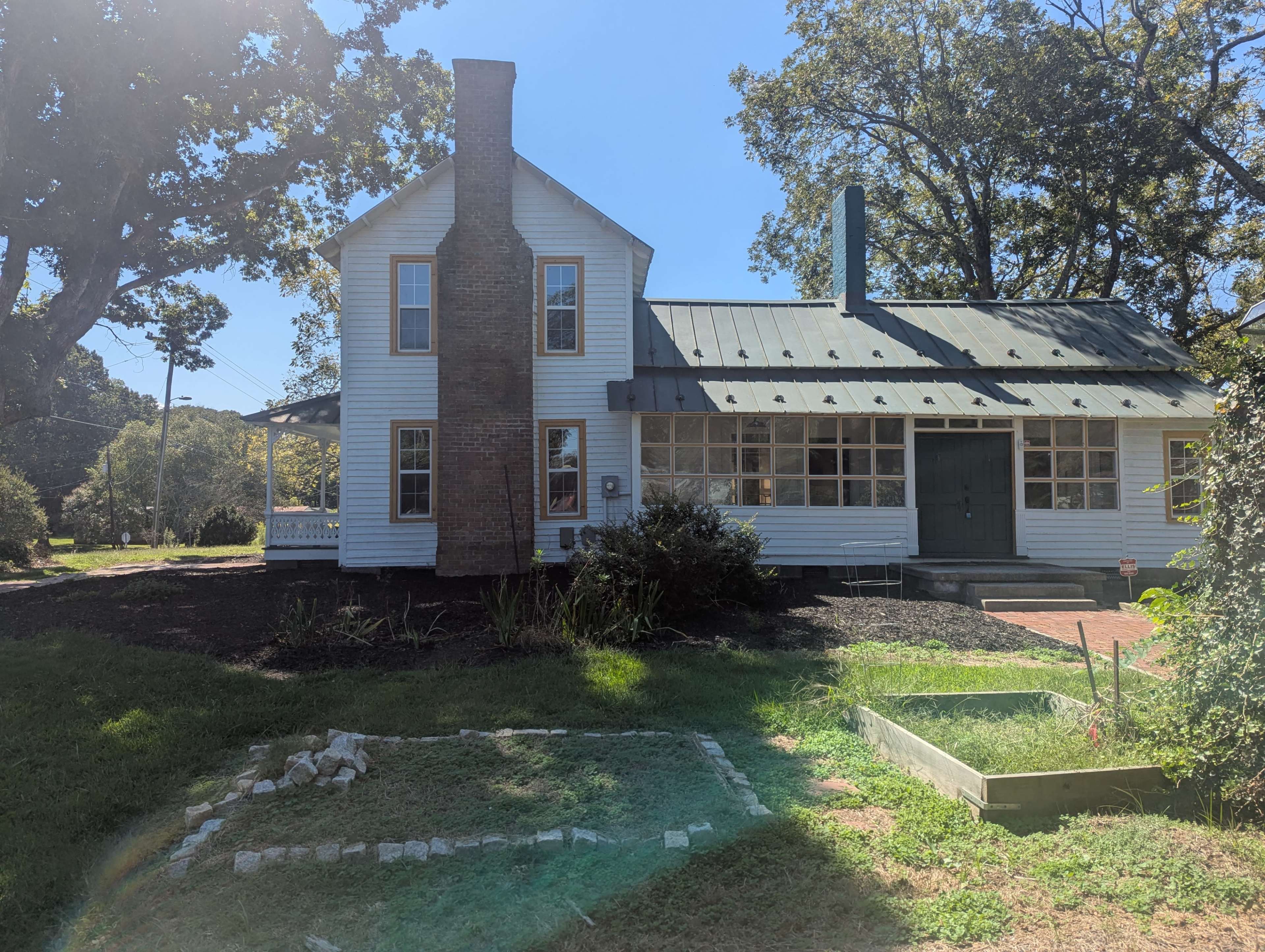 The image shows a two-story white house with a green metal roof, featuring a brick chimney and large windows, surrounded by grass and landscaping.