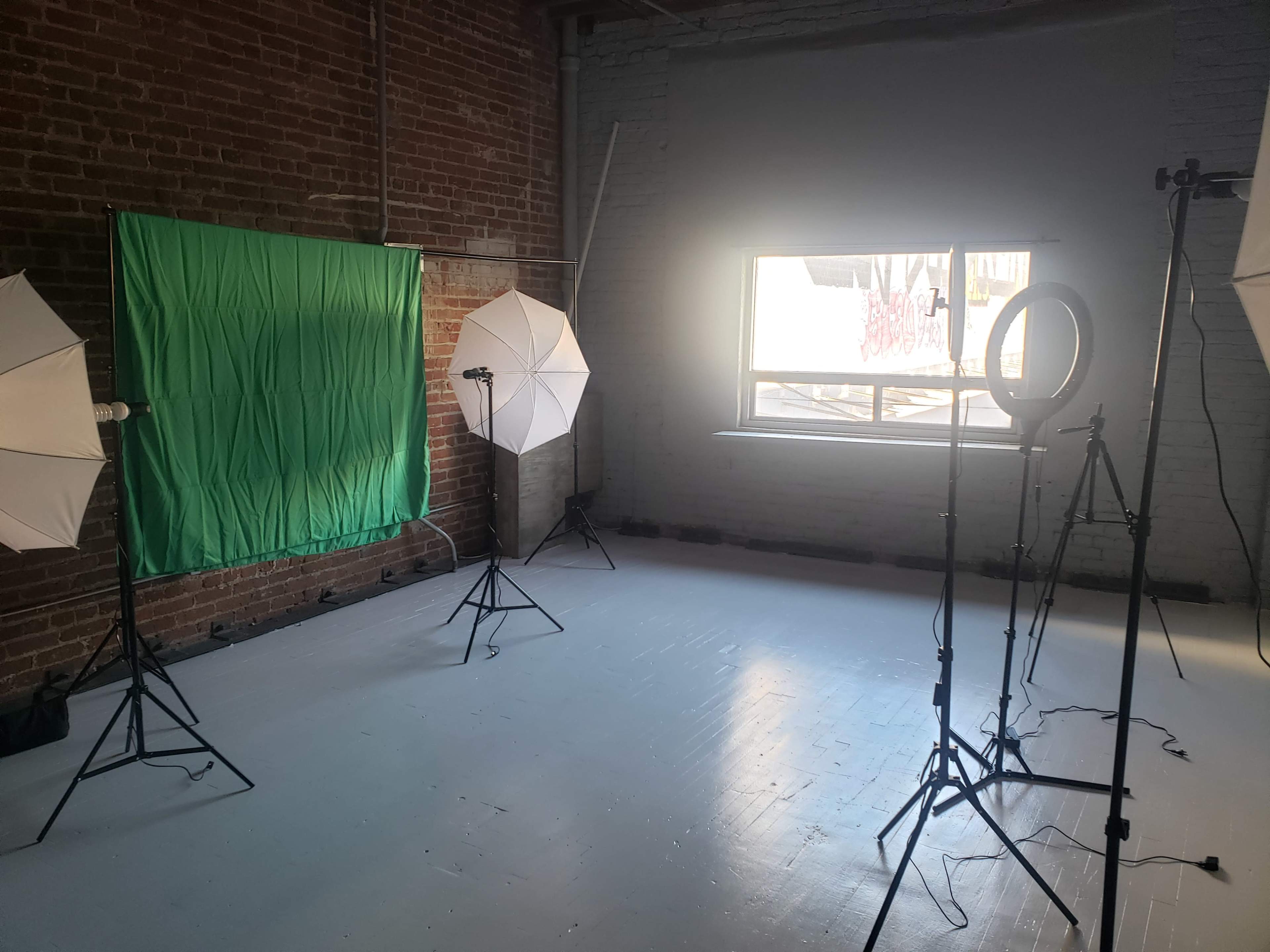 The image shows a photography studio with a green backdrop, two softbox lights, a ring light, and a window with graffiti visible outside.
