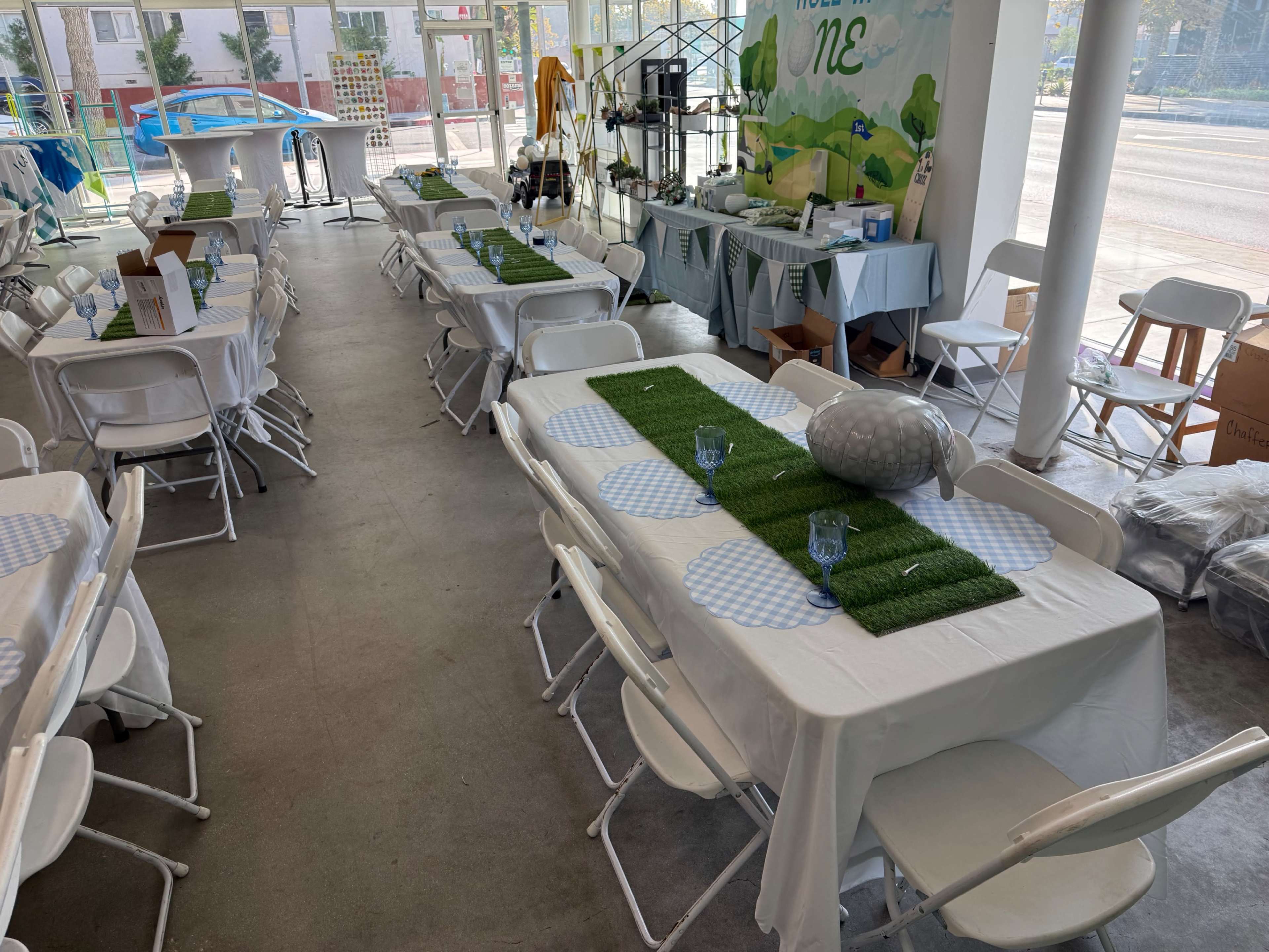 The image shows a spacious indoor area set up with several tables draped in white tablecloths, decorated with green and blue accents, ready for an event.