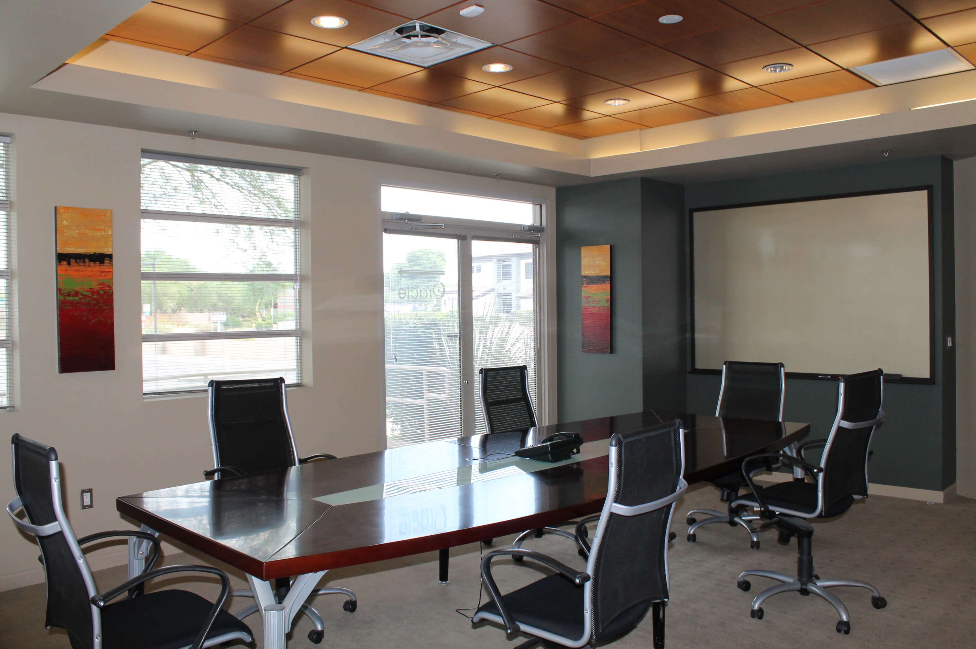 A conference room features a large rectangular table surrounded by six ergonomic chairs, with windows and a whiteboard on the walls.