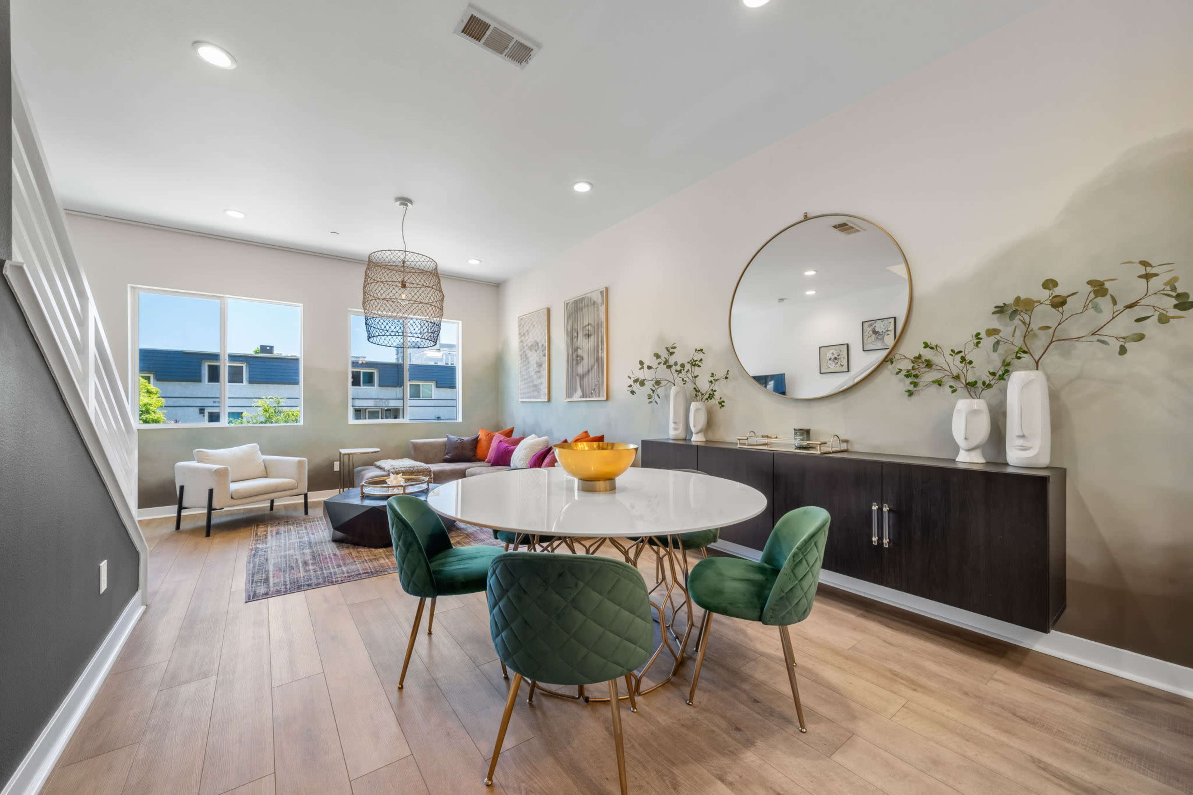 A modern dining area features a round table with green chairs, surrounded by a spacious living space with a sofa, decorative pillows, and large windows.