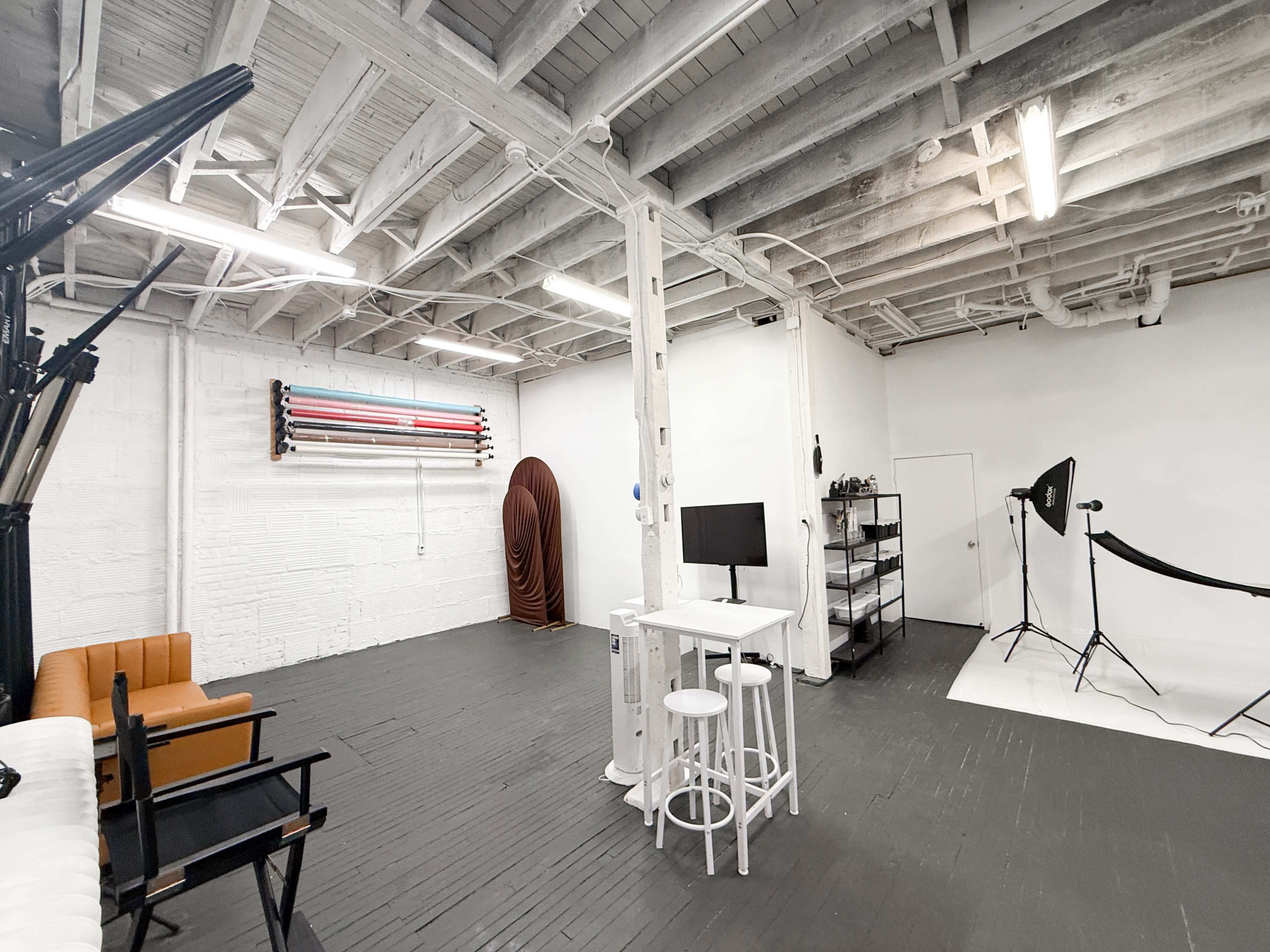 The image shows a spacious photo studio with a wooden floor, white walls, and various equipment arranged, including a backdrop stand and light setup.