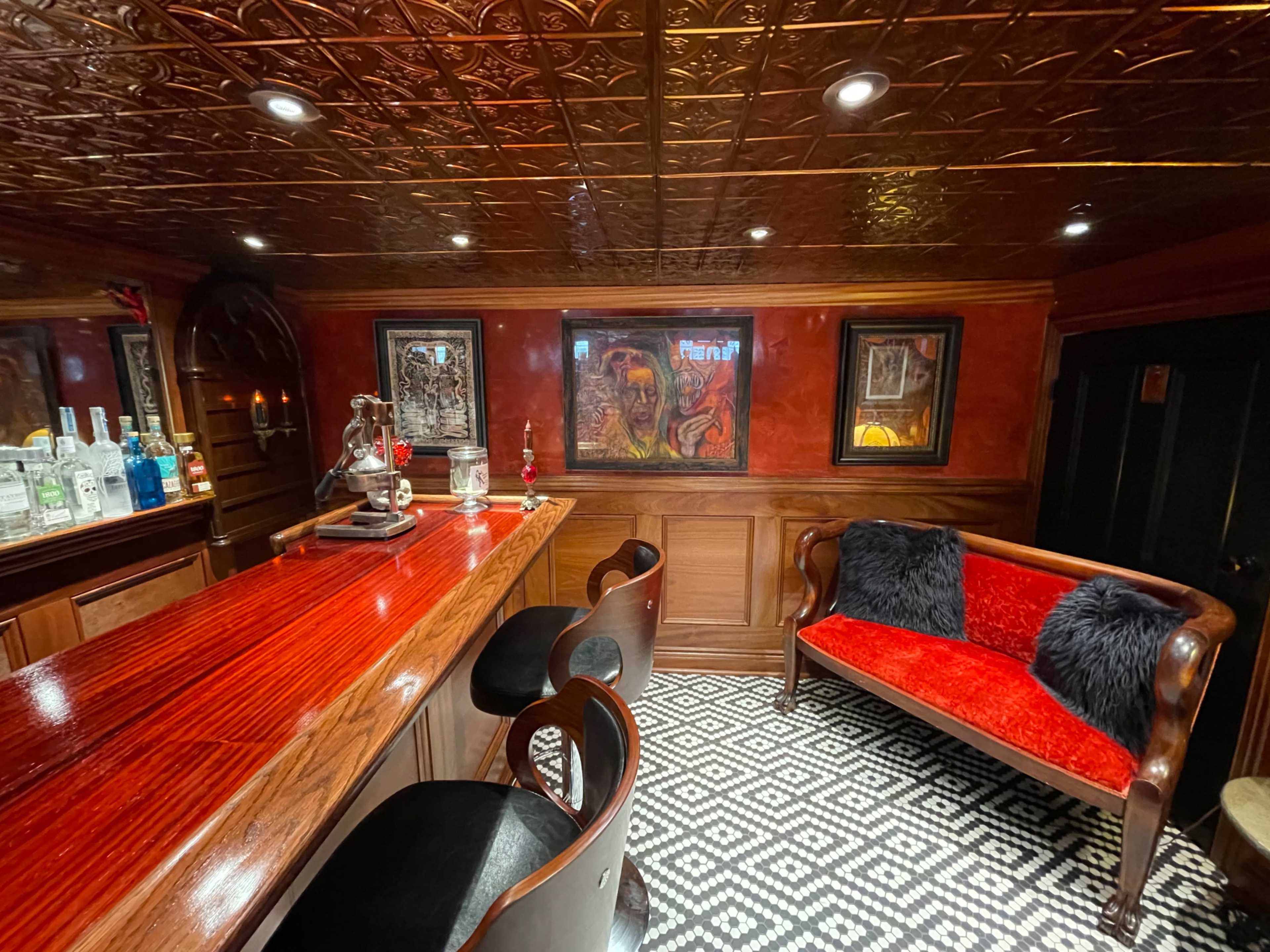 The image shows a dimly lit bar area with a shiny wooden countertop, black chairs, and red accent details, featuring artwork on the walls and a patterned rug on the floor.