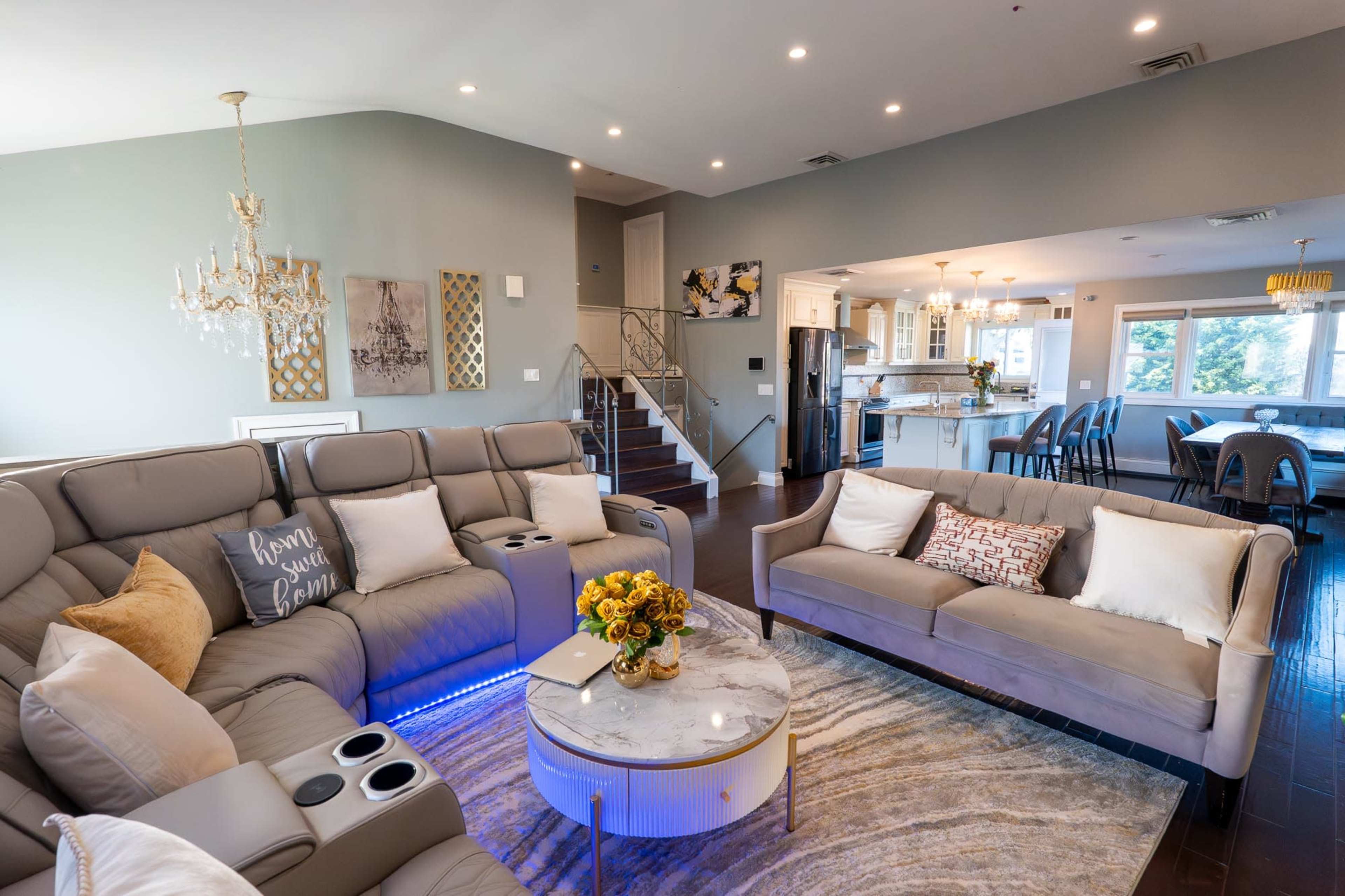 The image shows a modern living room with a sectional sofa, a round coffee table, and a well-lit open-concept layout that connects to a dining area and kitchen.