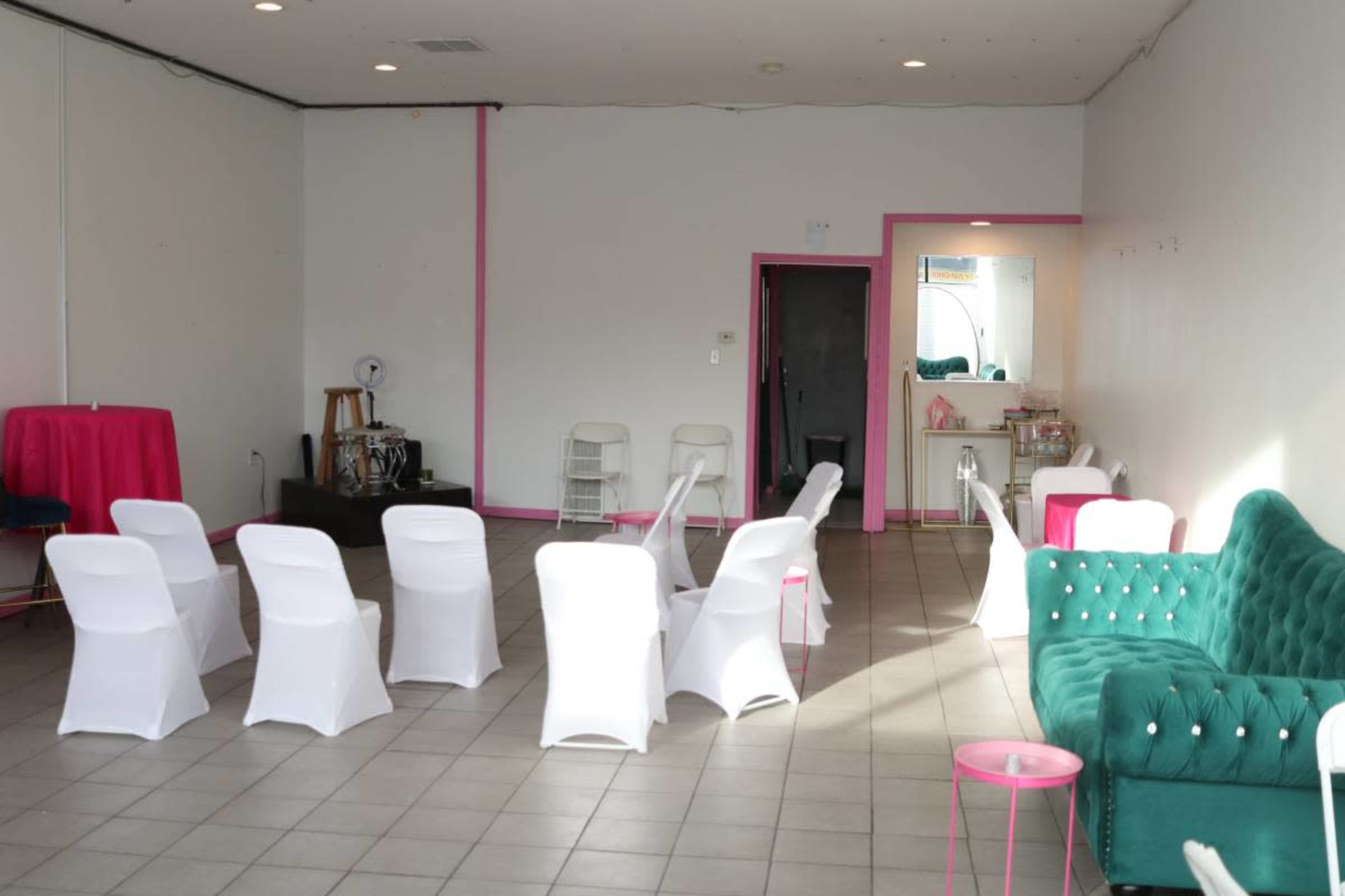 The image depicts a spacious, brightly lit room with white walls, arranged with several white chairs facing a small table and a green tufted sofa.