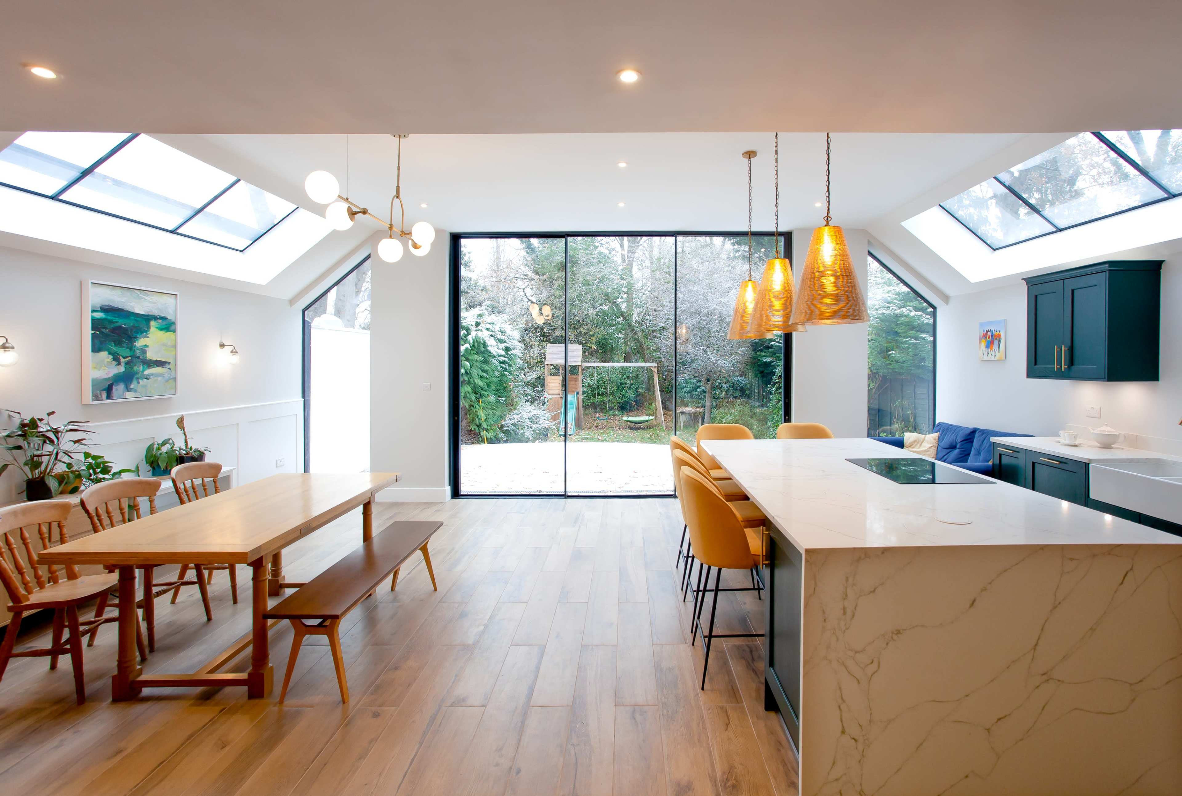 A modern kitchen and dining area features a large marble island, wooden dining table, and glass doors leading to an outdoor space.