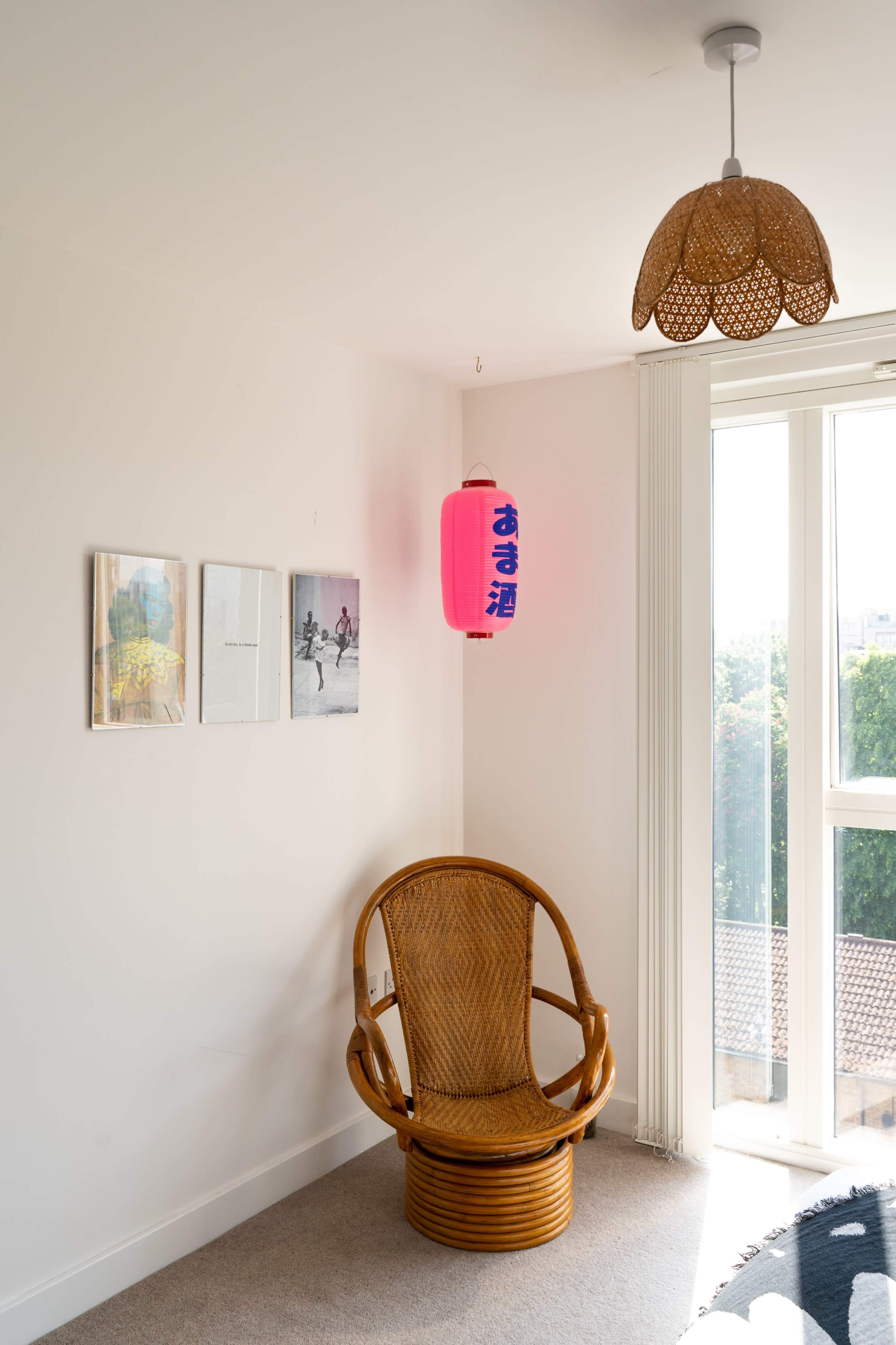 A corner of a bright room with a rattan chair, a woven pendant light, and a pink lantern hanging on the wall beside framed artwork.