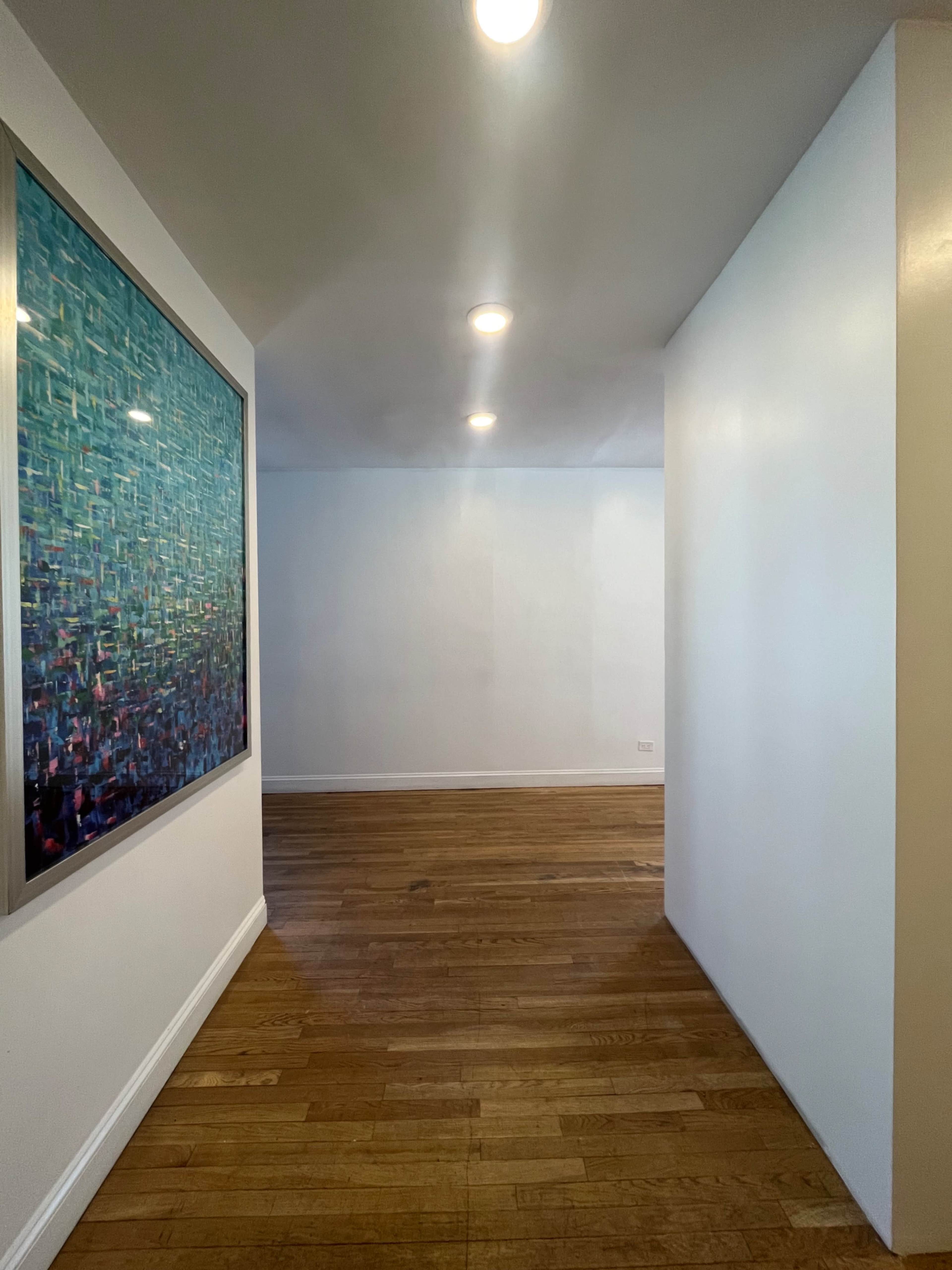 The image shows a spacious hallway with wooden flooring, featuring a large abstract painting on the left wall under bright ceiling lights.