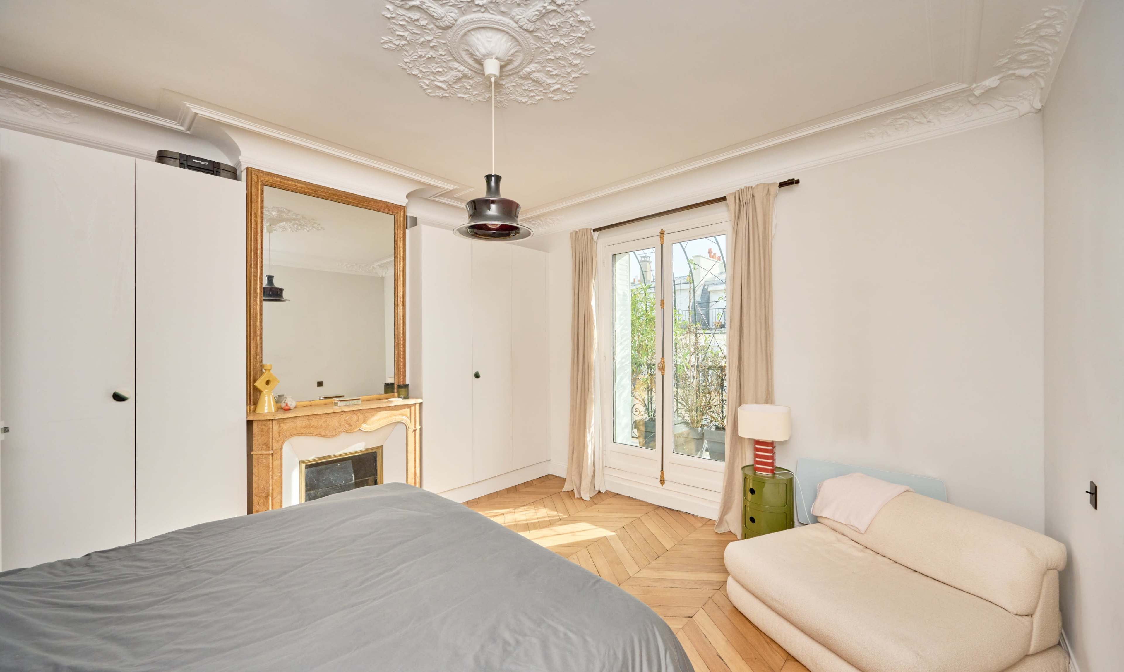 The image shows a bright bedroom featuring a bed, a small sofa, a decorative mirror, and large windows with sheer curtains.