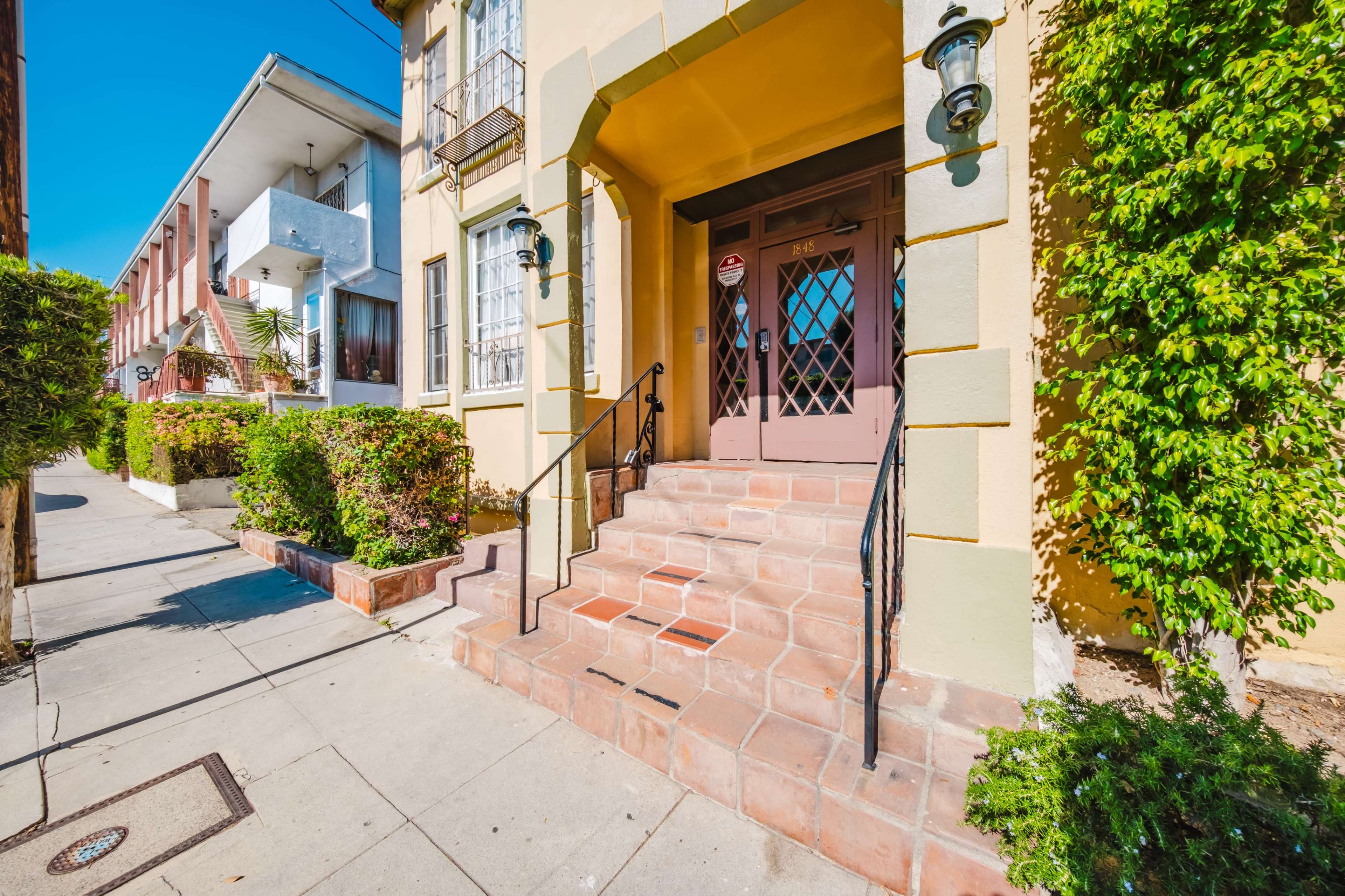 The image shows a building entrance with a set of steps leading up to a wooden door, flanked by wall-mounted lanterns and surrounded by neatly trimmed shrubs.