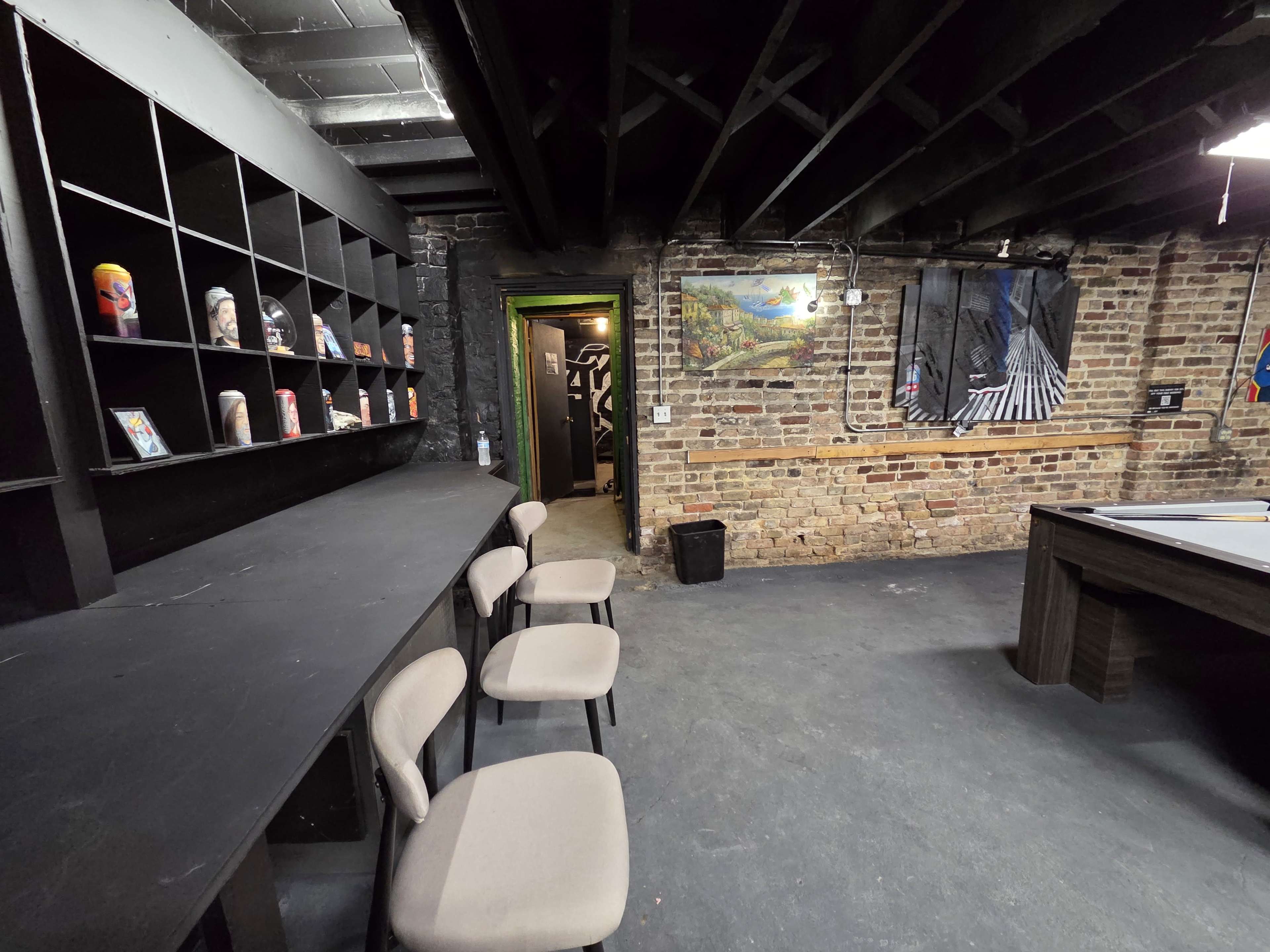 The image shows a dimly lit room with exposed brick walls, a long black shelf displaying various items, and a few light-colored chairs arranged in front of it.