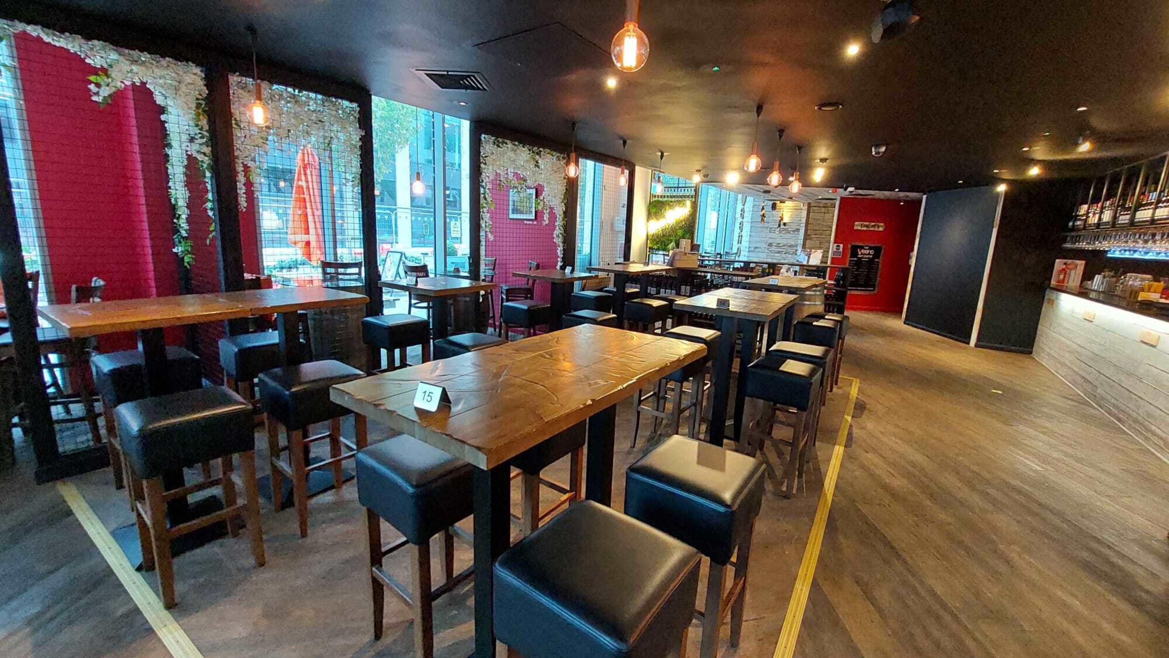 A spacious interior of a restaurant or bar with wooden tables and black leather chairs, illuminated by pendant lights.