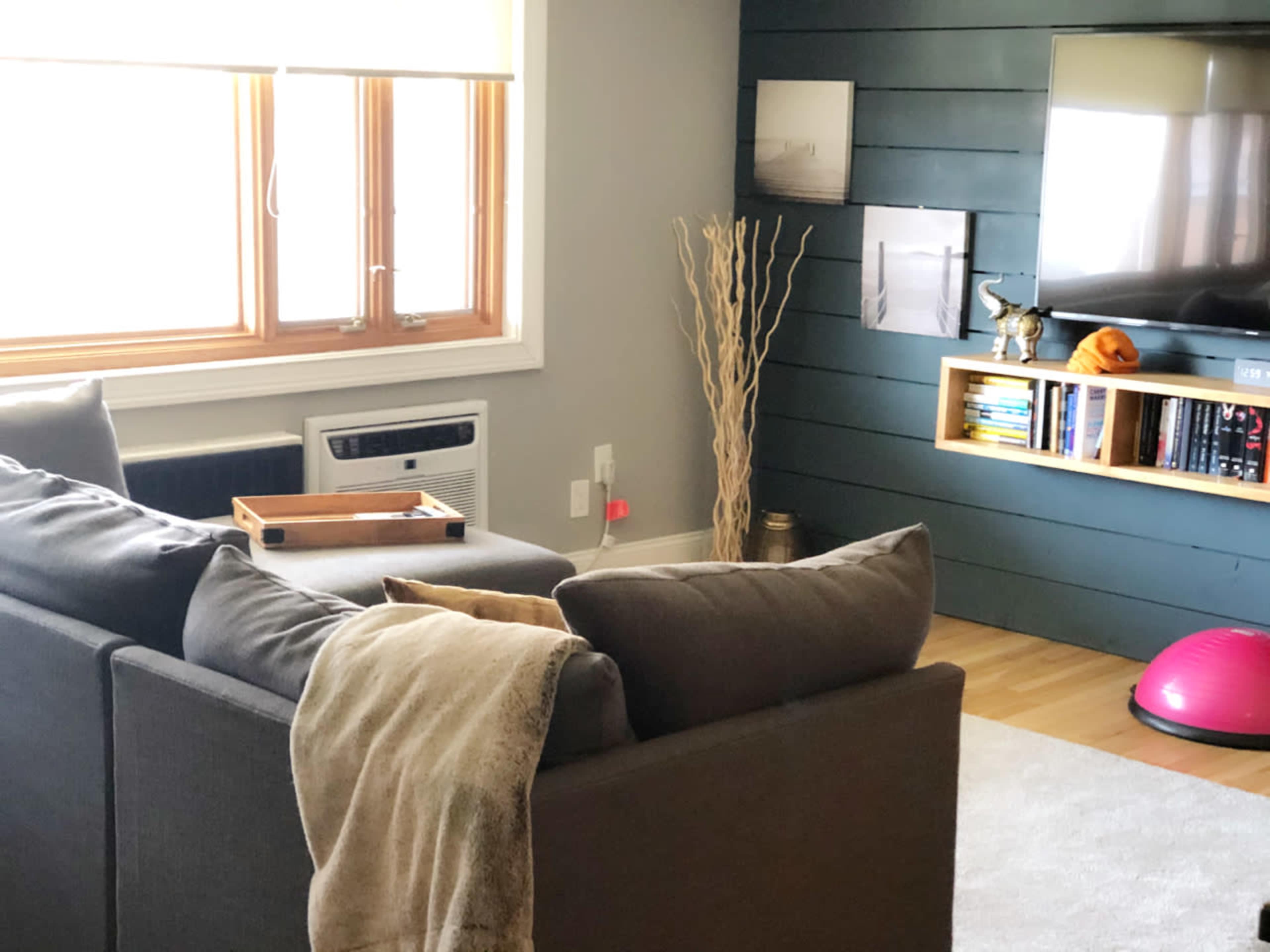 A living room features a gray sofa with cushions, an air conditioning unit, a television mounted on a dark wall, and shelves with books and decorative items.
