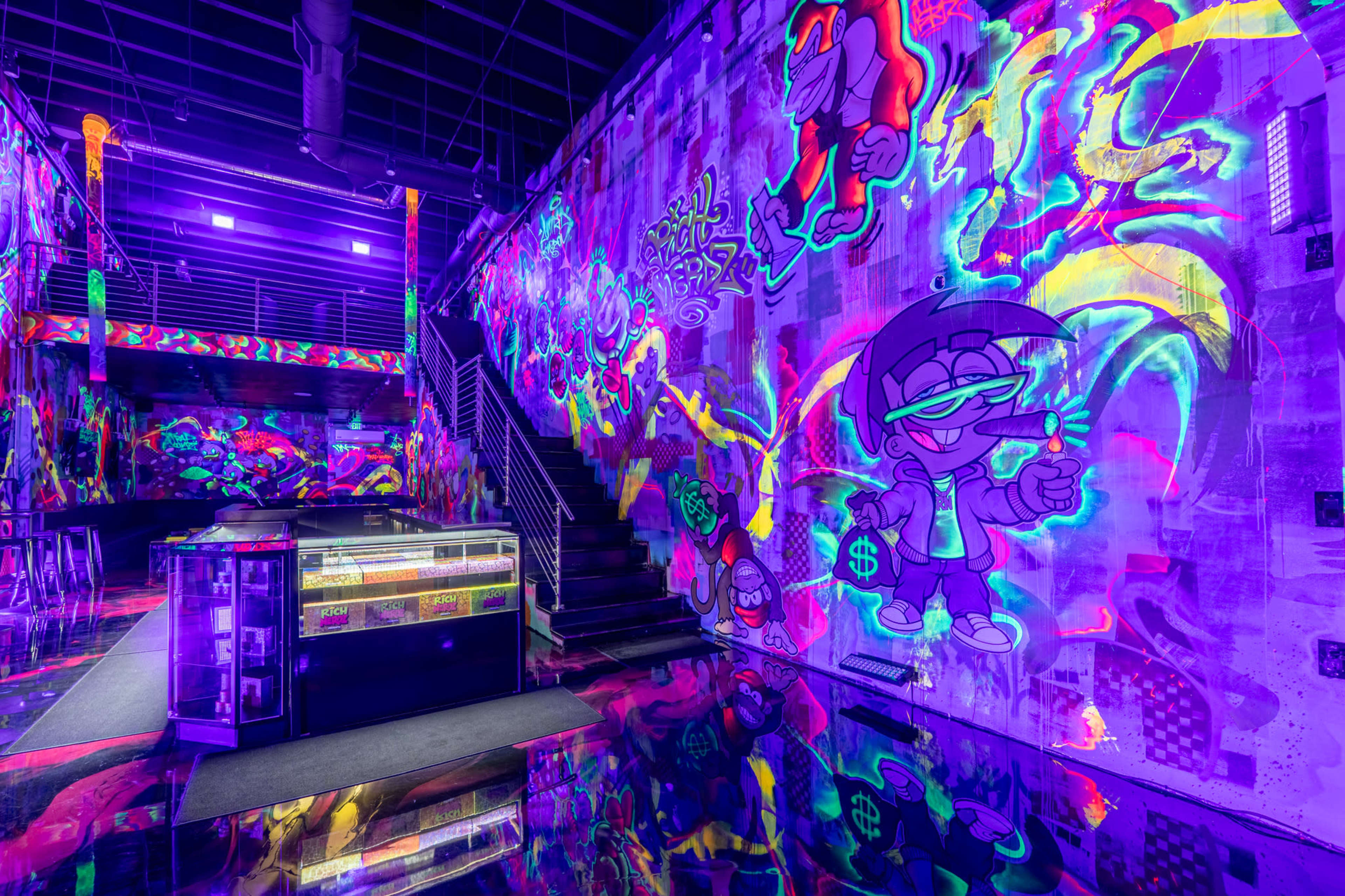 The image shows a brightly lit indoor space featuring colorful graffiti on the walls, with a reflective floor and a visible staircase leading to an upper level.