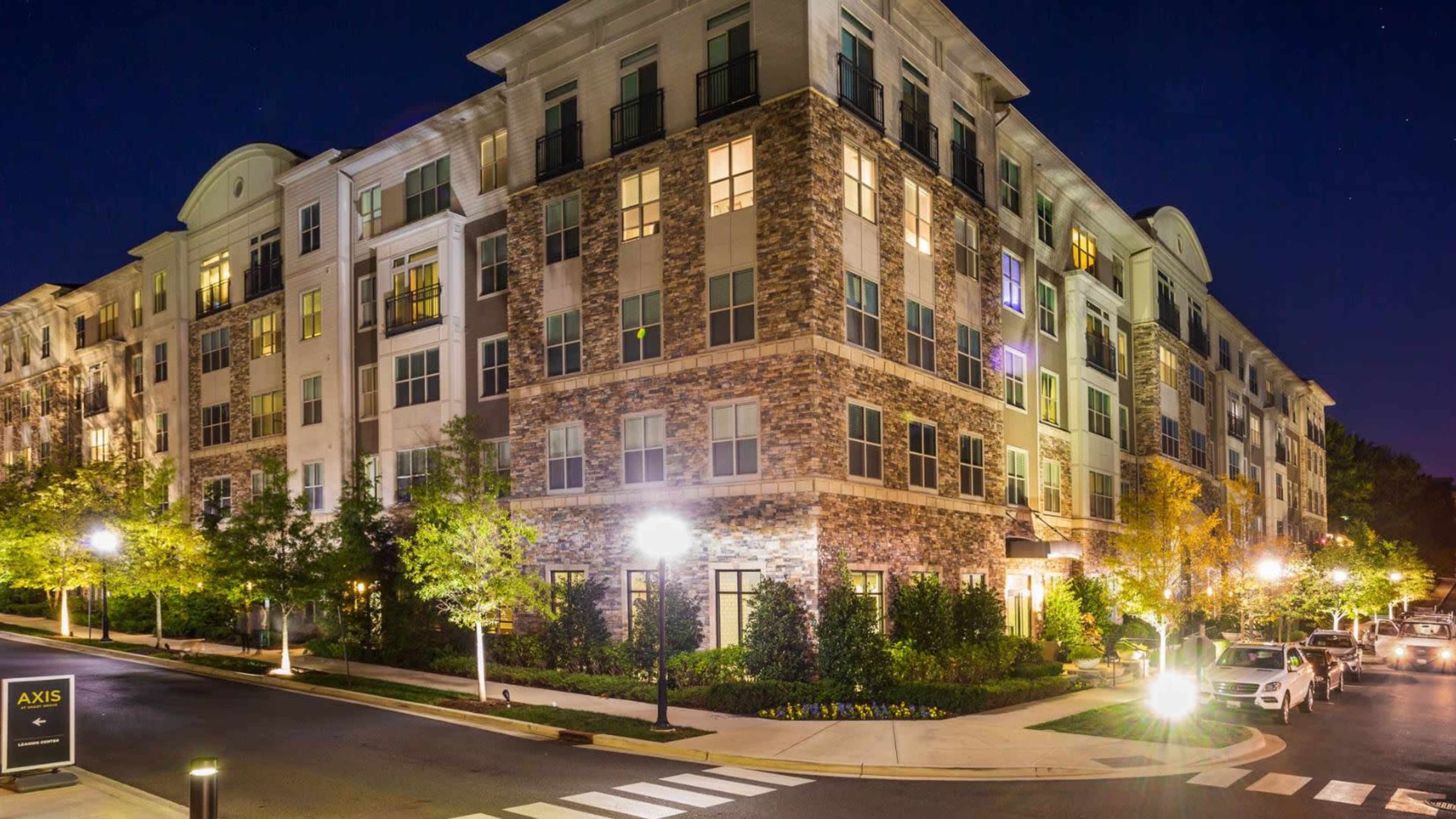 A modern apartment building is illuminated at night, showcasing its stone facade and landscaped surroundings.