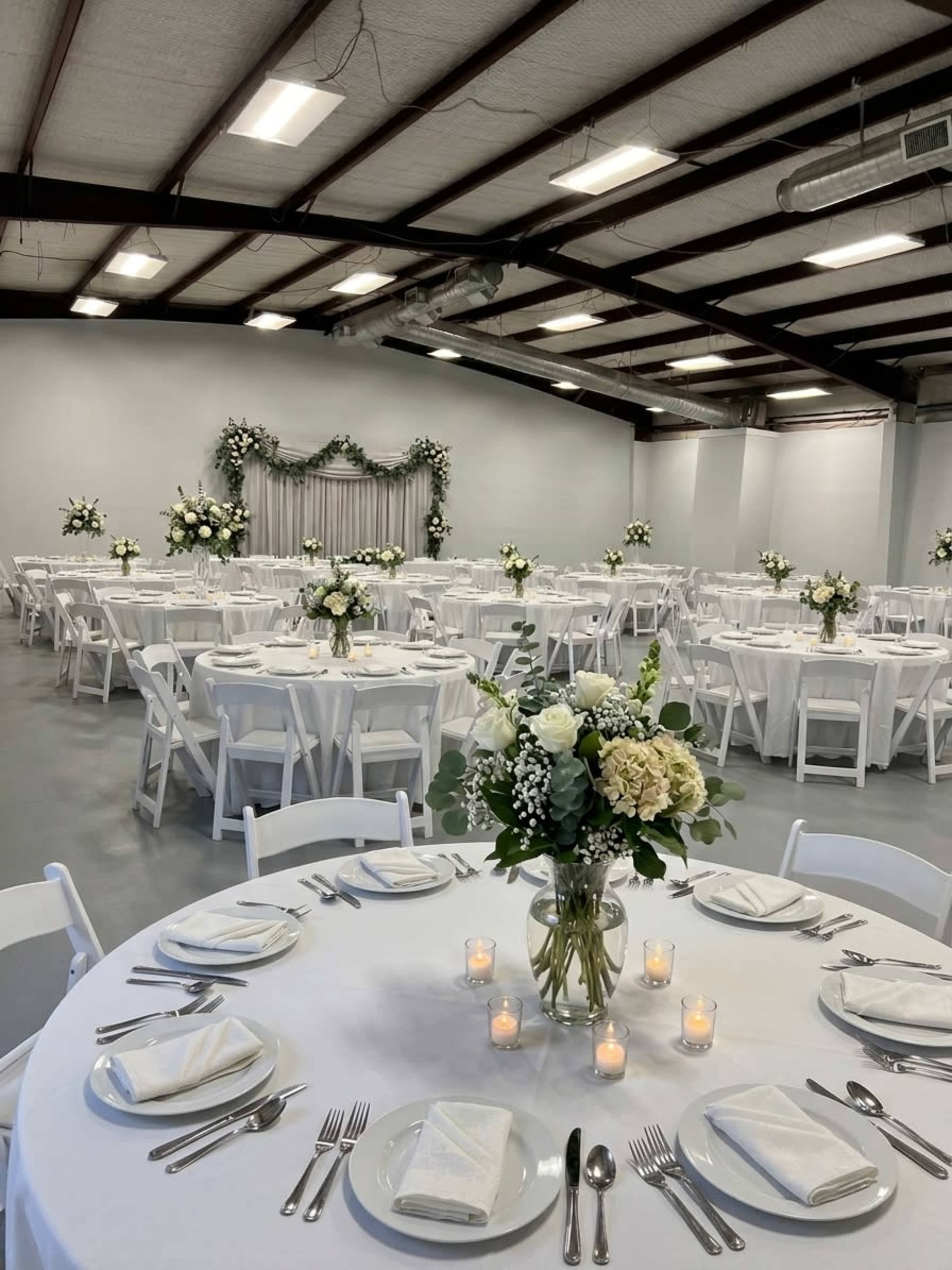 The image shows a spacious event hall set up for a formal gathering, featuring elegantly arranged round tables with centerpieces and white folding chairs.