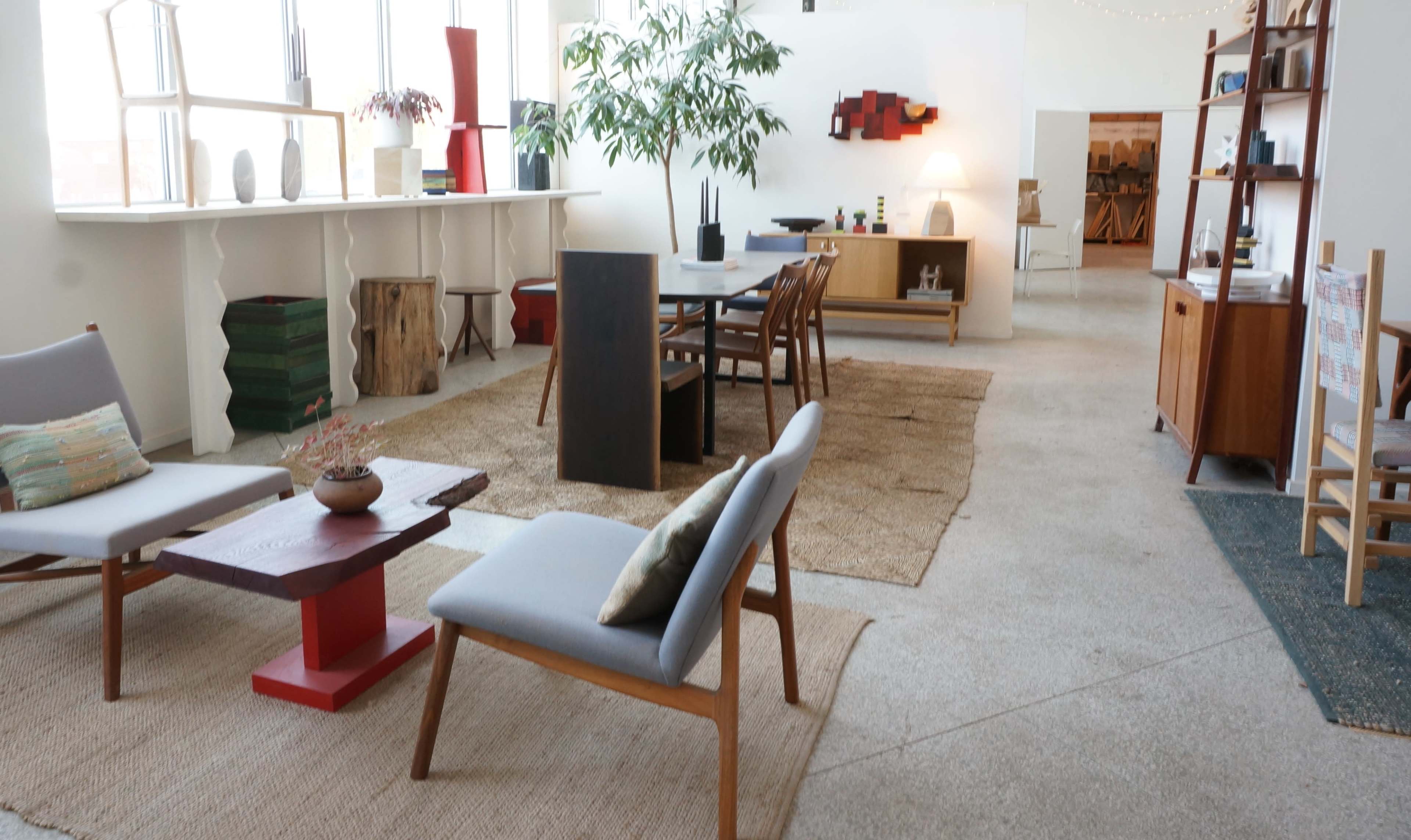 The image shows a spacious, well-lit interior with modern furniture, including a wooden dining table, chairs, and a pair of armchairs arranged on a textured area rug.