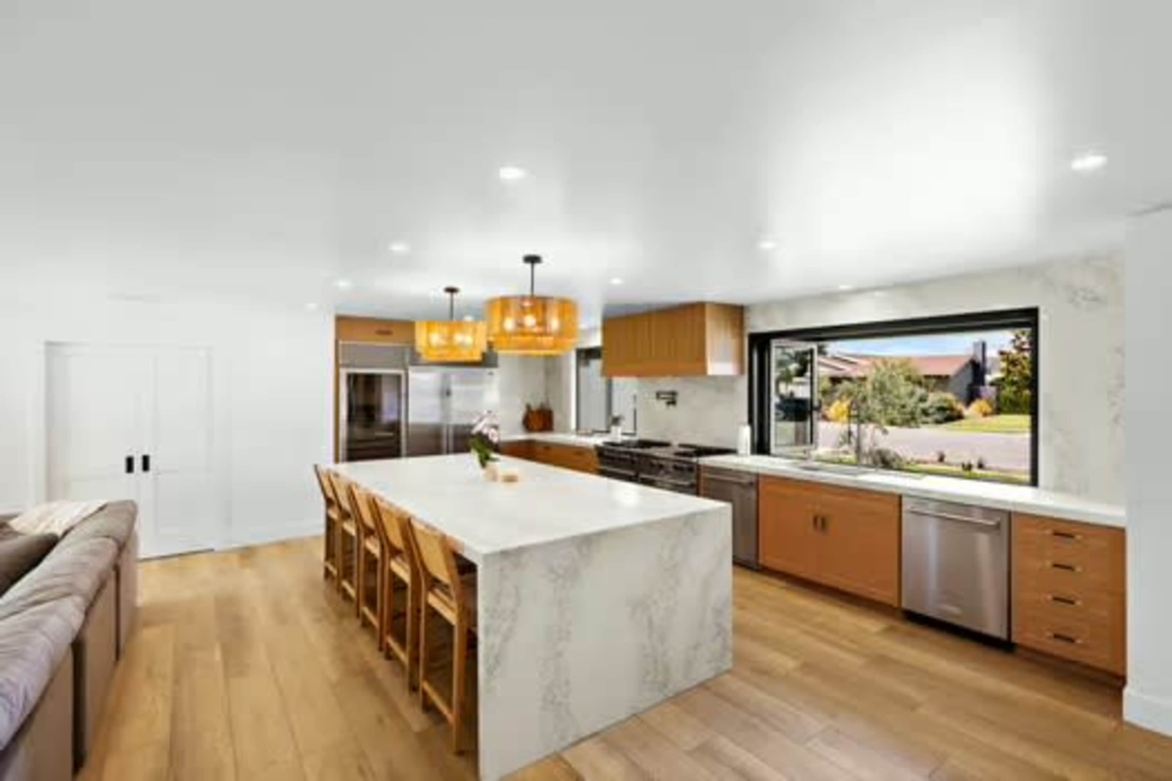 The image shows a modern kitchen with a large island, wooden cabinetry, and large windows overlooking an outdoor view.