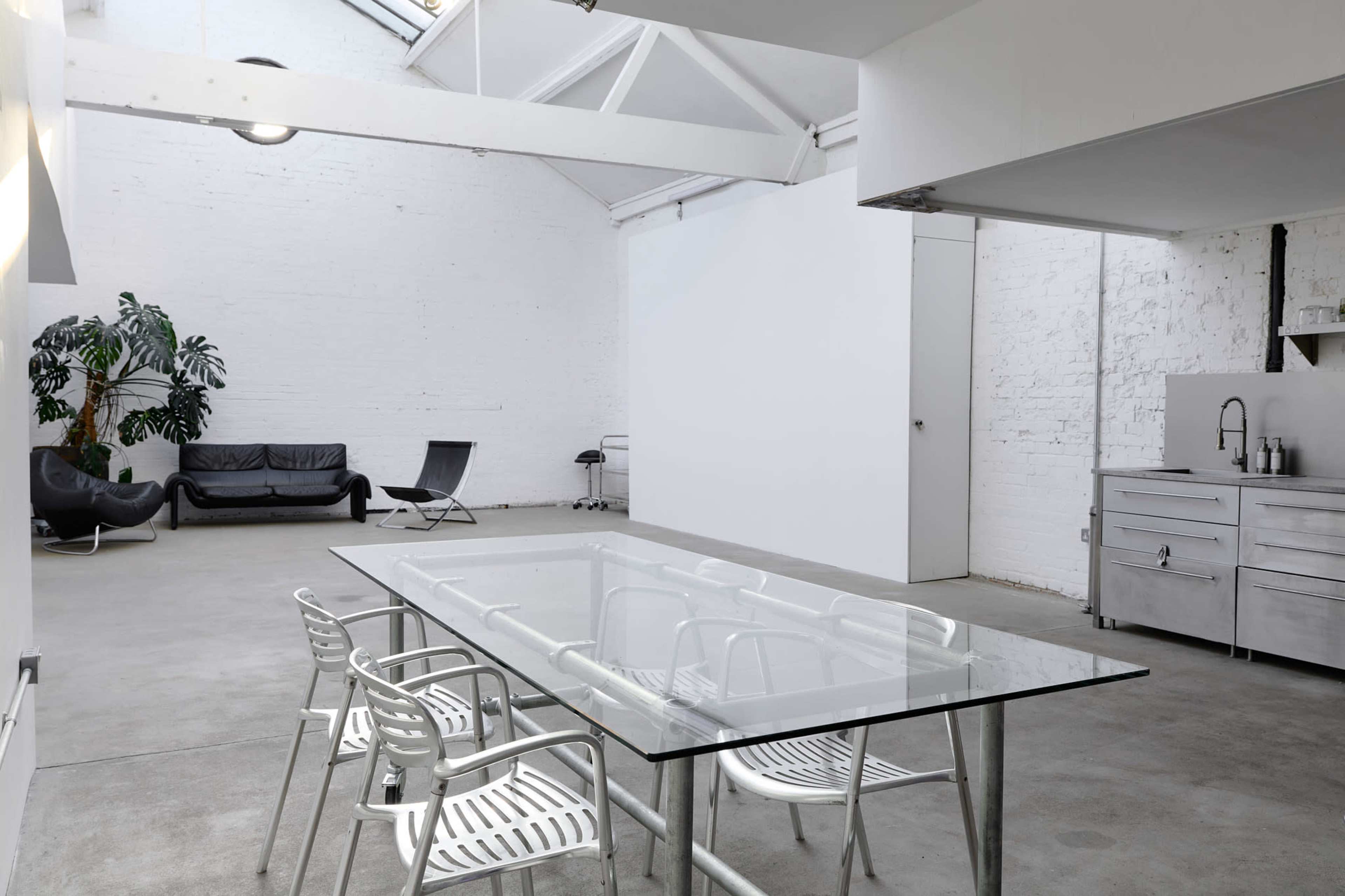 The image shows a spacious, modern interior with a glass dining table surrounded by metal chairs, a black couch, and large windows allowing natural light to enter.