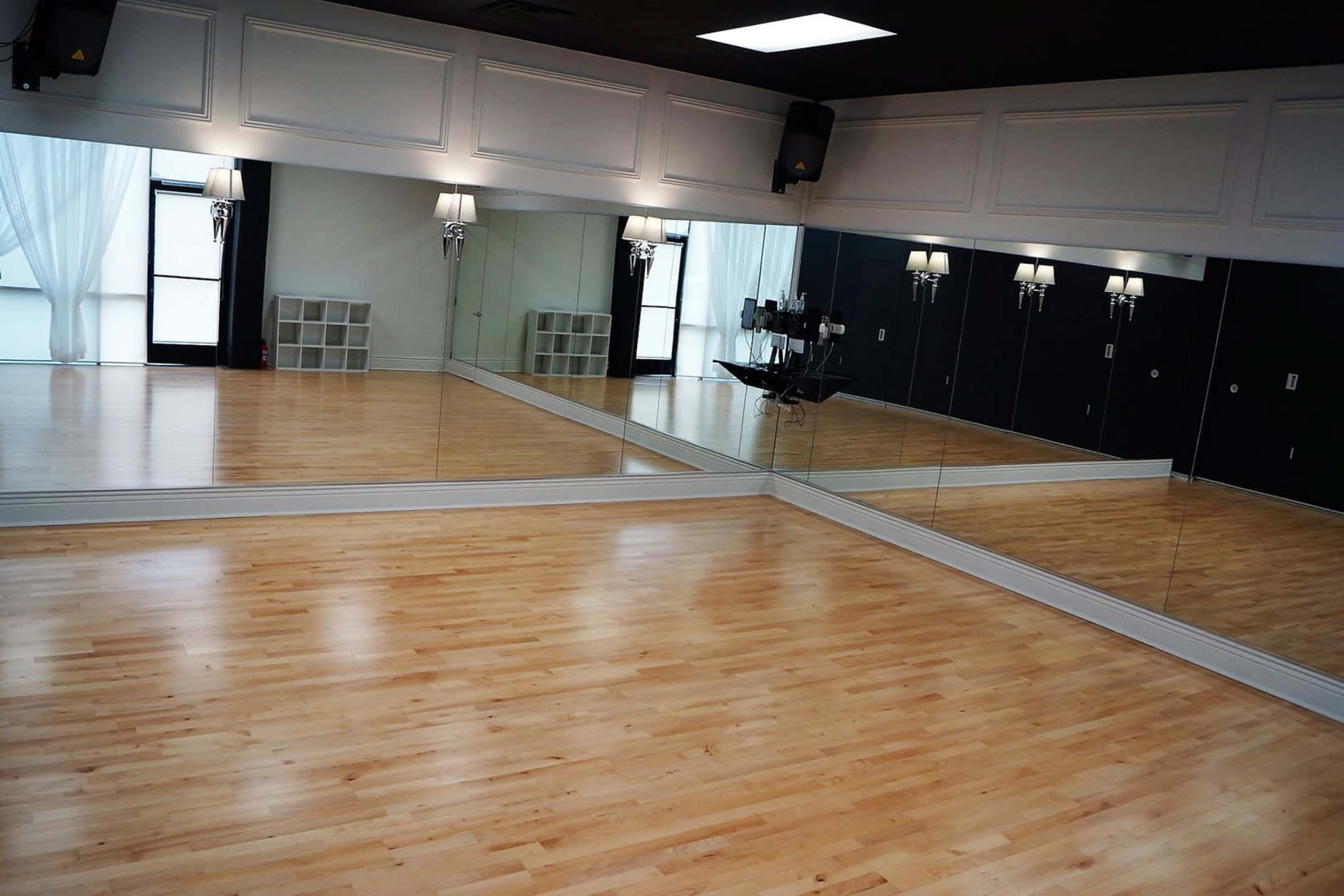 An empty dance studio with mirrored walls and polished wooden floors, illuminated by wall sconces and large windows.