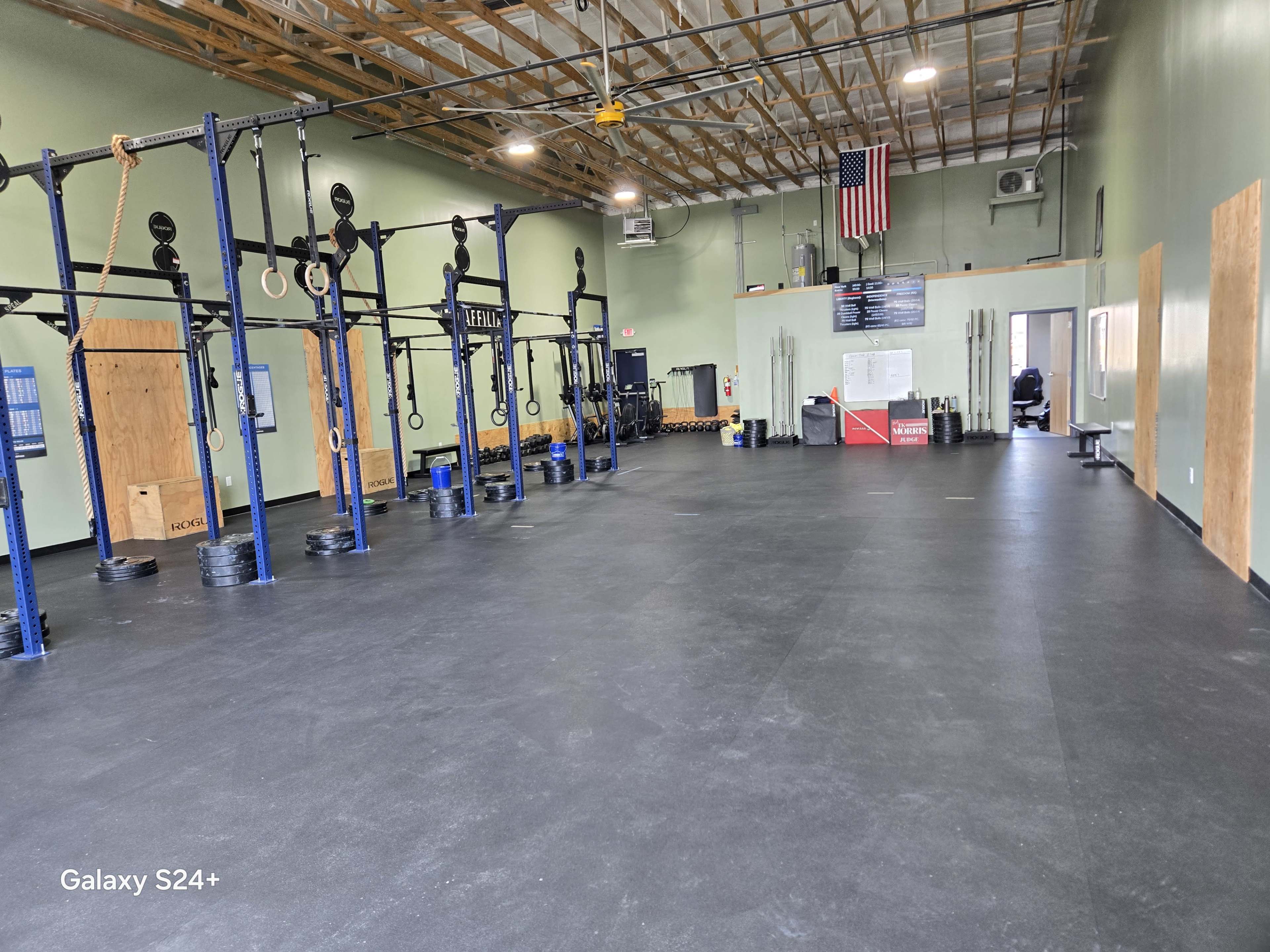 The image shows a spacious indoor gym area with exercise equipment, including pull-up bars and weightlifting platforms, along with a flag hanging on the wall.