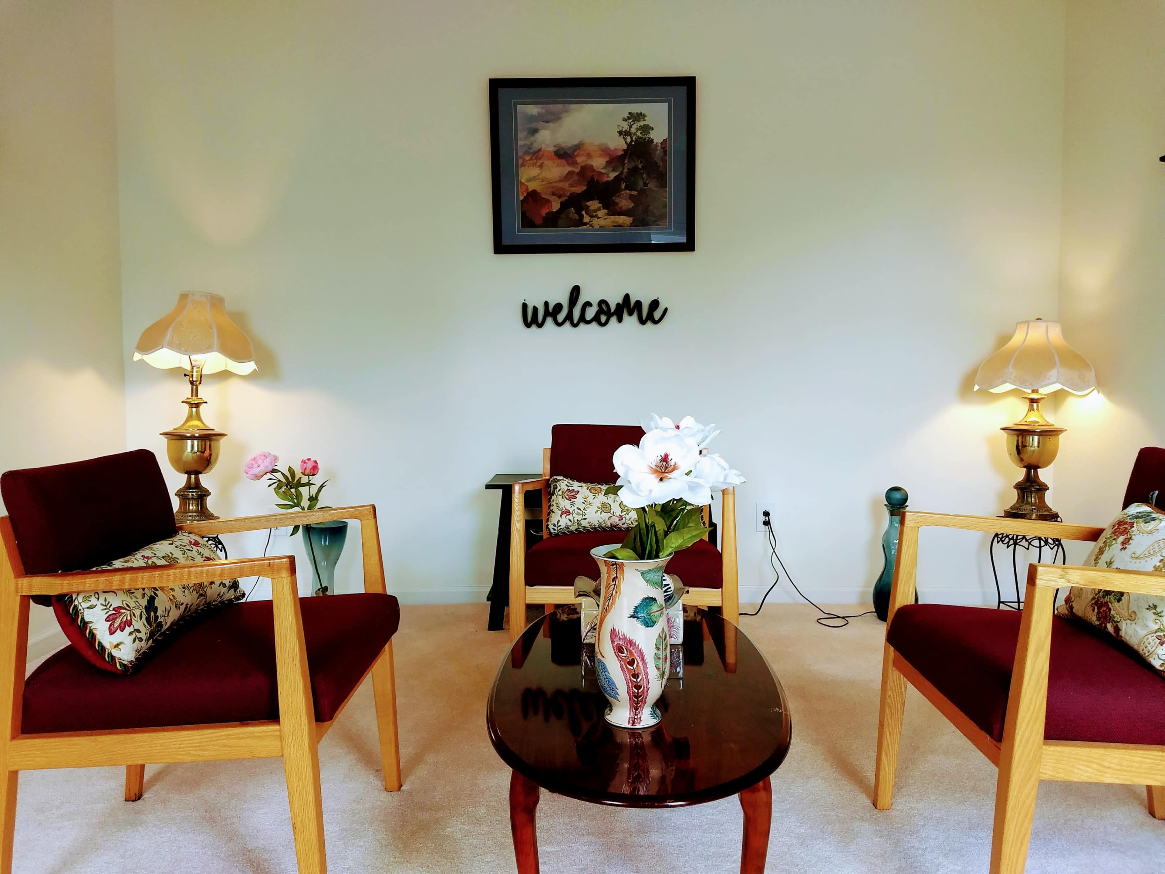 A cozy sitting area with four wooden chairs, a round table, and two lamps, accompanied by a welcome sign and floral decorations.