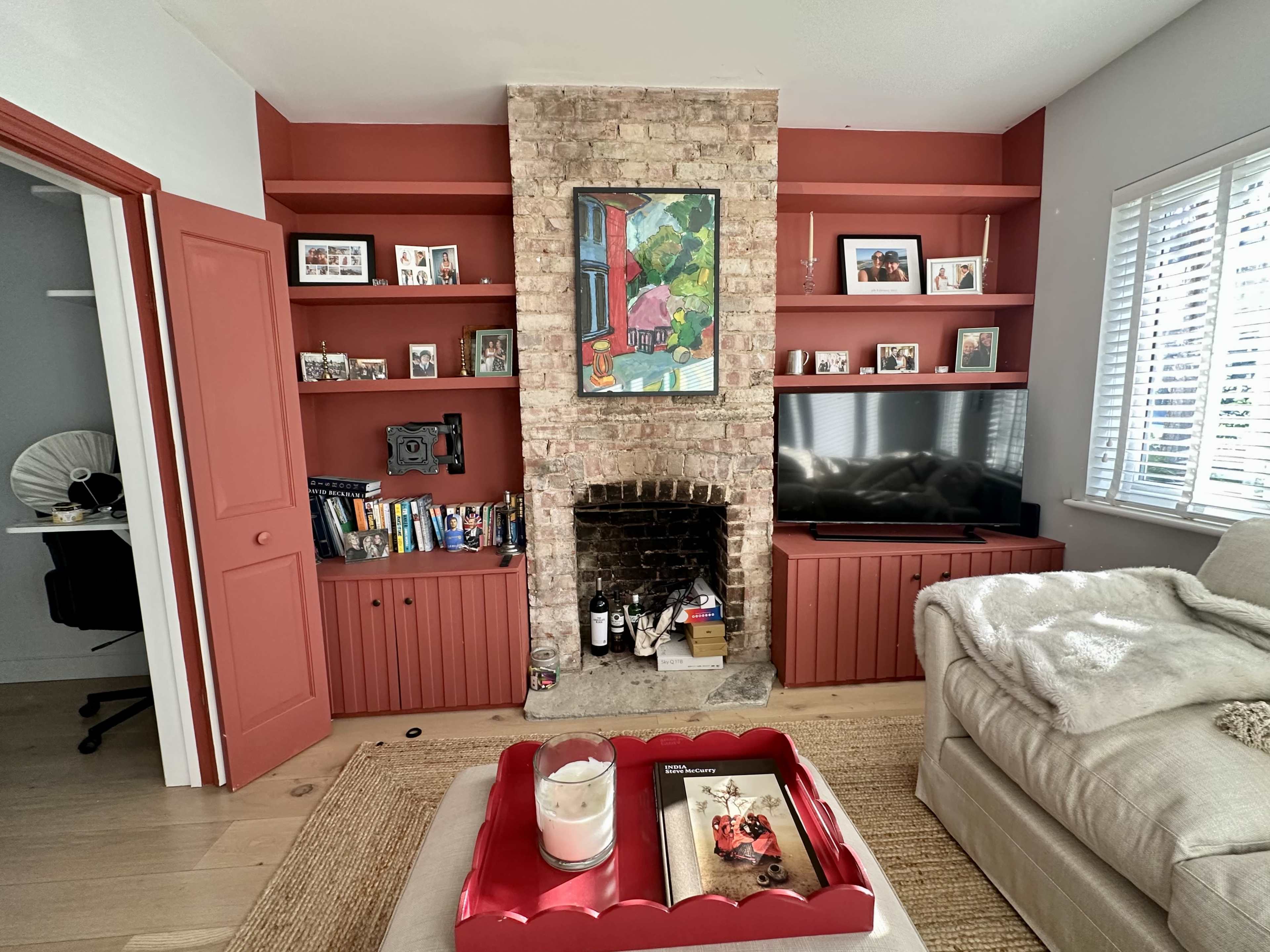 The image shows a cozy living room featuring a brick fireplace, built-in shelves filled with photographs and books, and a television set against a red backdrop.