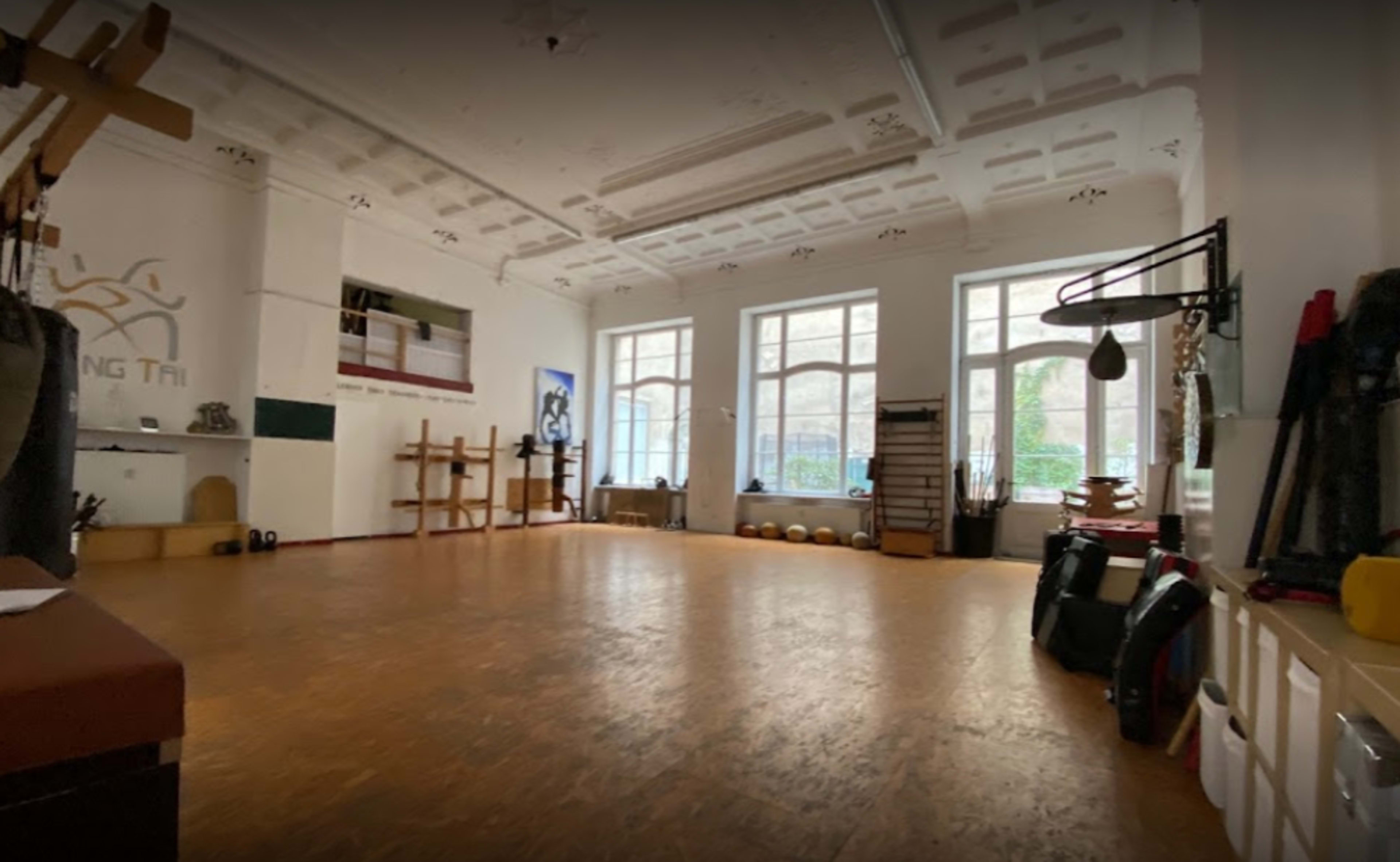 The image shows a training studio with wooden floors, large windows, and various martial arts equipment neatly arranged.