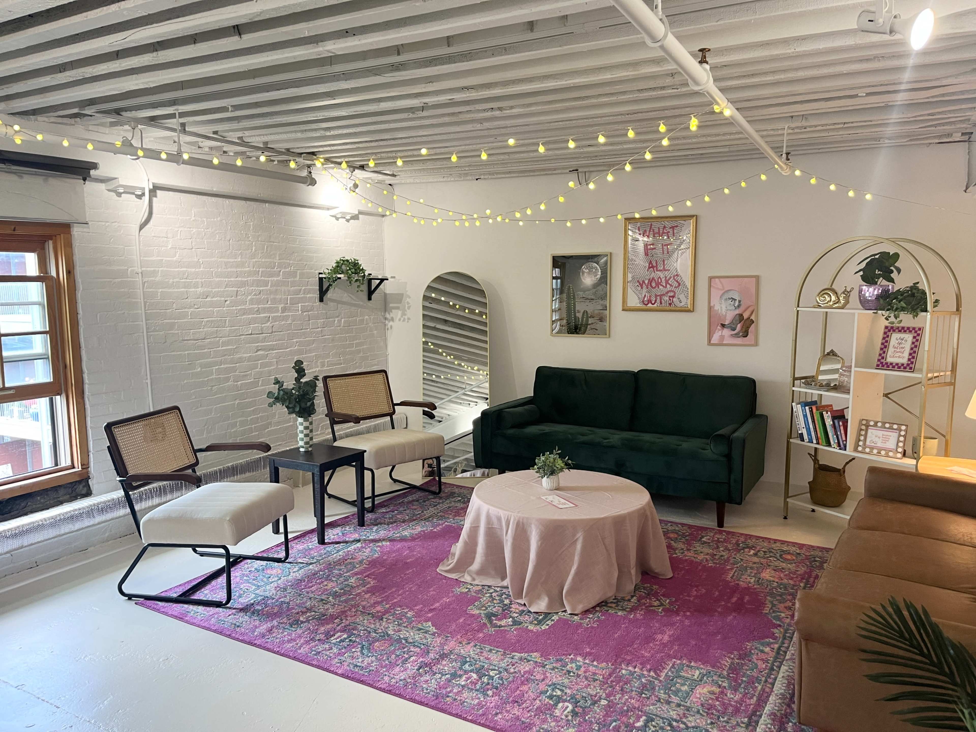 A modern living space with a green sofa, two chairs, a round coffee table on a pink rug, and a bookshelf surrounded by warm lighting.
