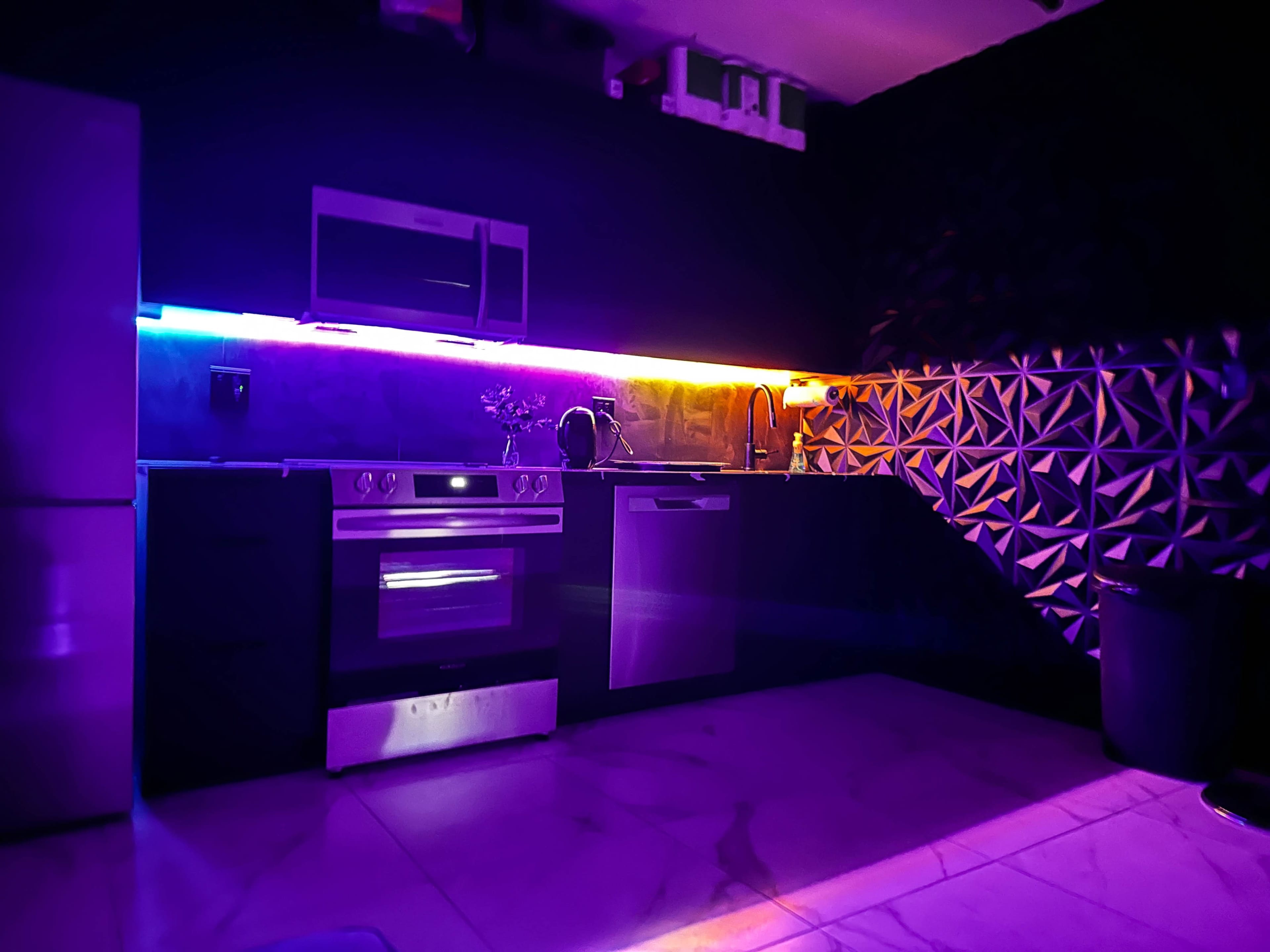 The kitchen features sleek black cabinets, stainless steel appliances, and colorful LED lighting illuminating the countertop and backsplash.