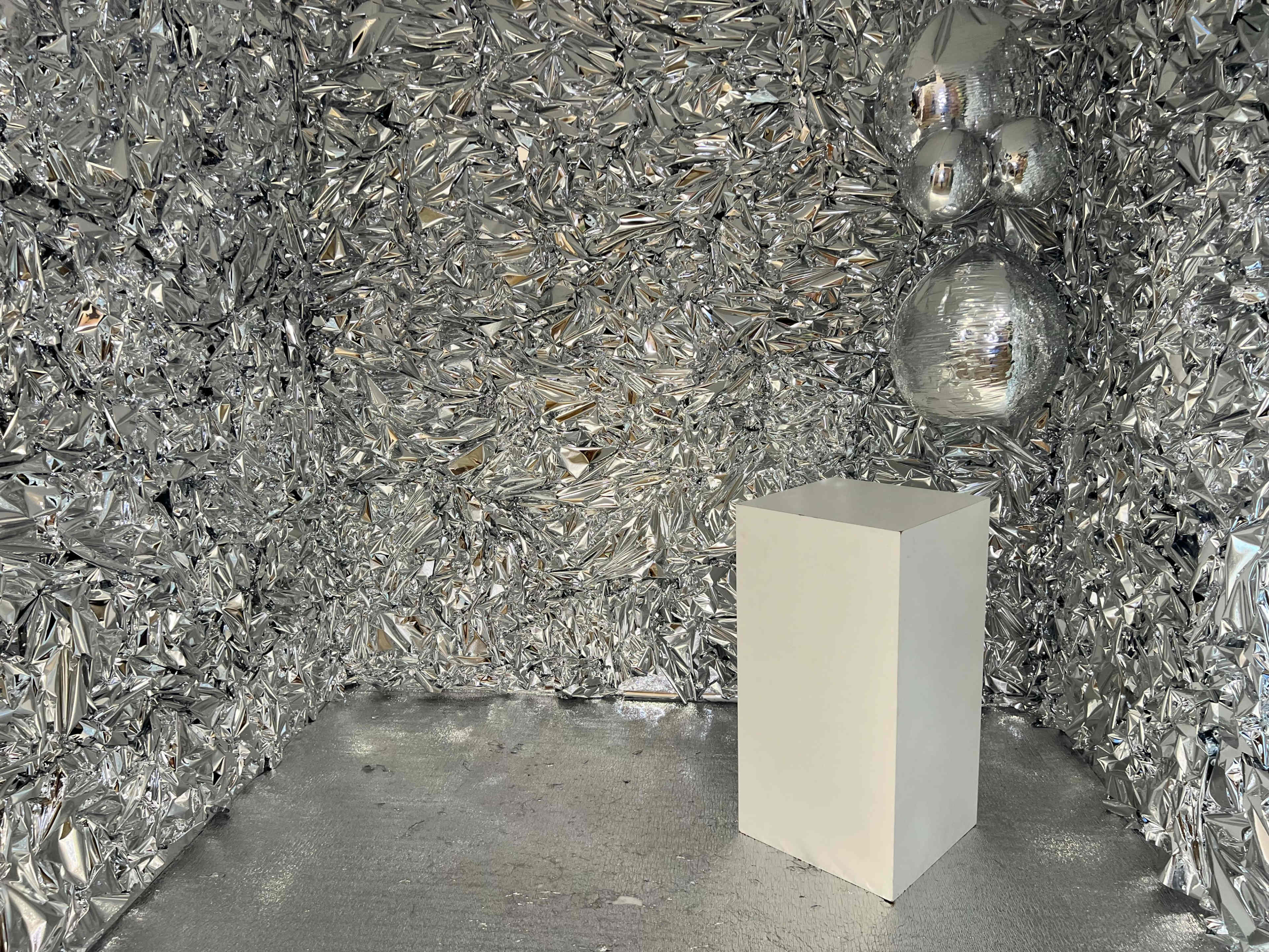 A white pedestal stands in the center of a room entirely lined with crumpled silver foil.