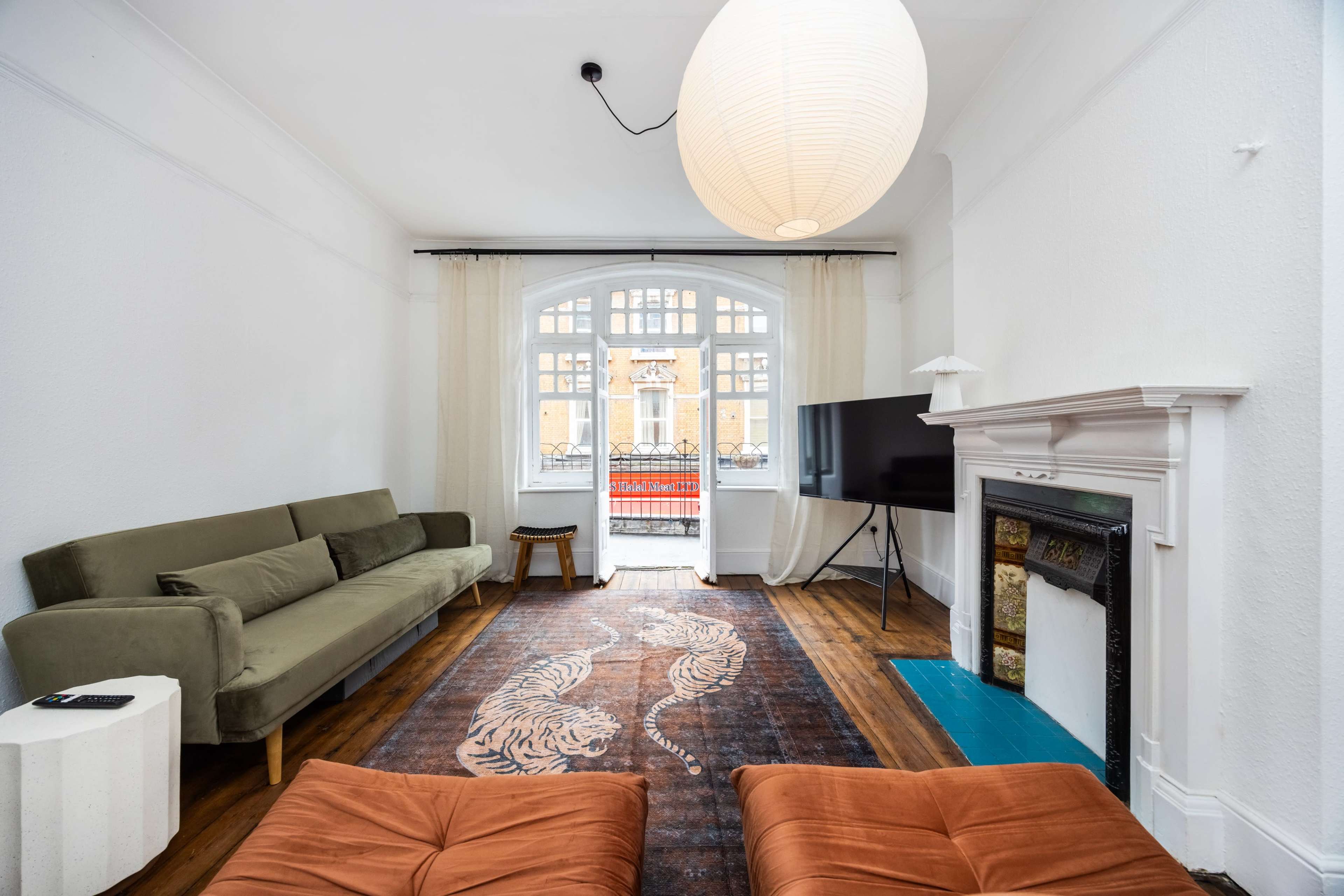 The living room features a green sofa, two brown ottomans, a television, and a large window that opens to a view outside.