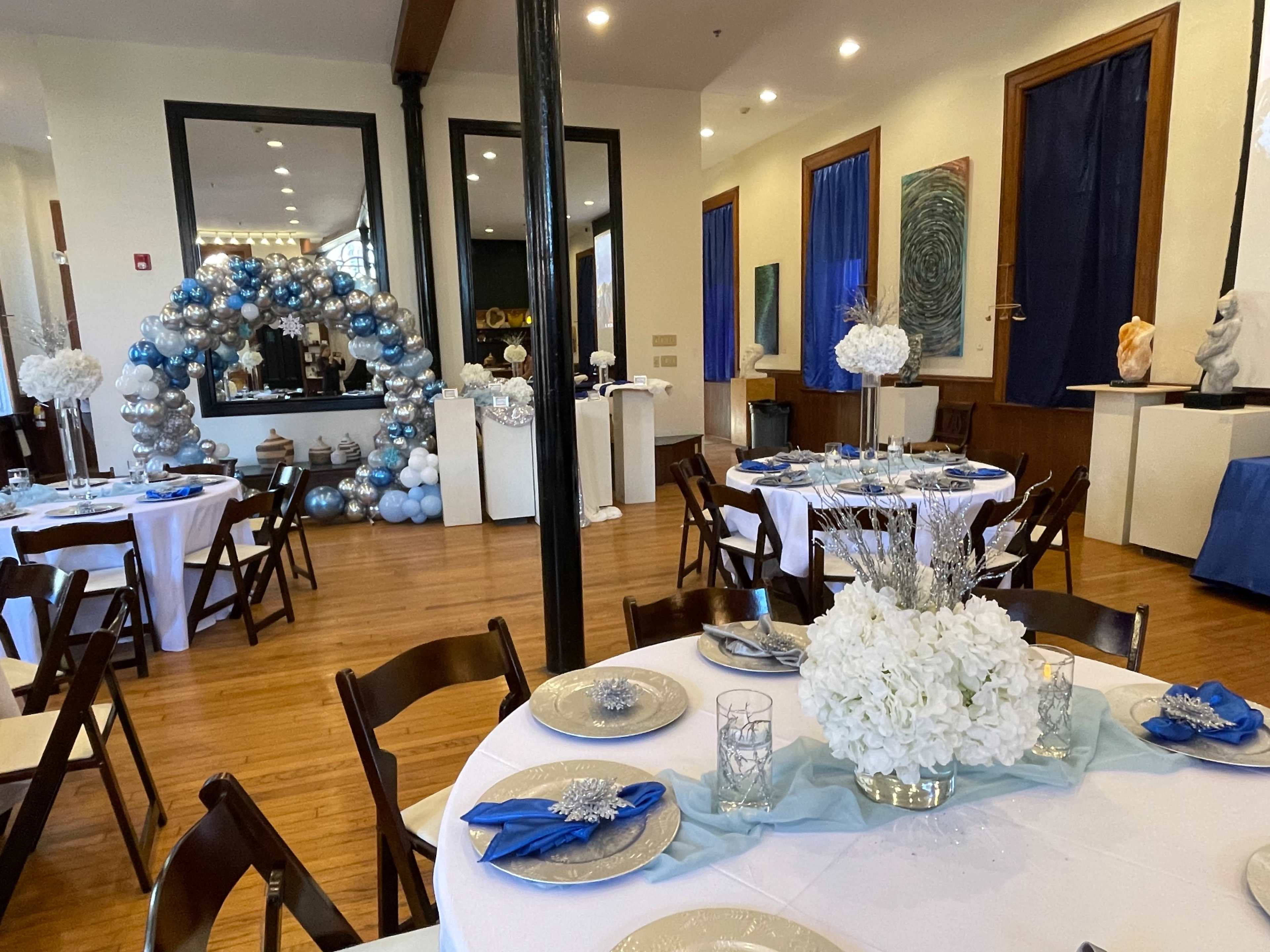 The image shows a decorated event space with tables set for a gathering, featuring blue and white decor, an arch of balloons in the background, and artworks on the walls.