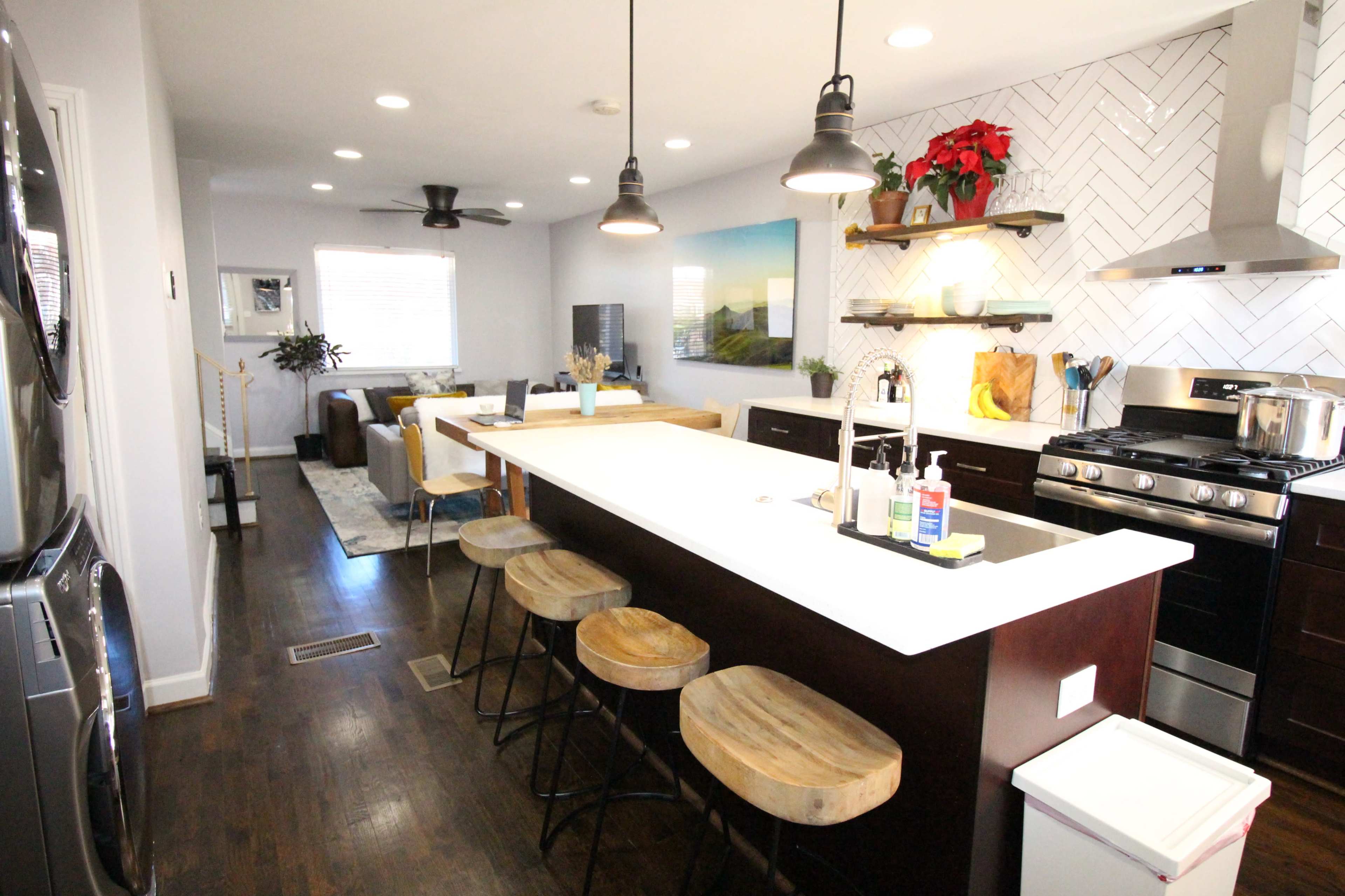 A modern kitchen with a large island, wooden bar stools, and a herringbone backsplash, illuminated by pendant lights.