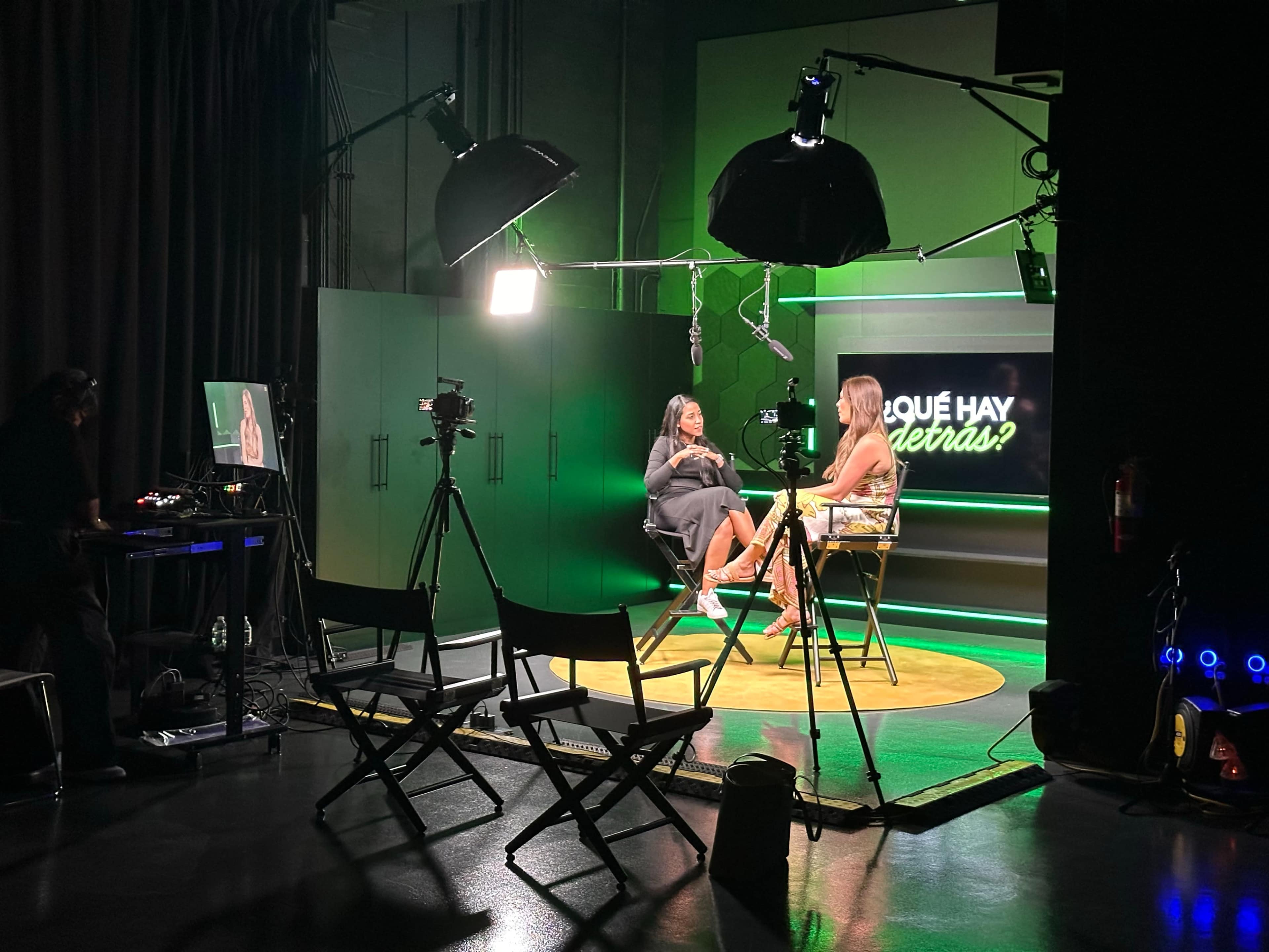The image shows a television studio setup with two seated hosts engaged in a conversation under bright lights, while cameras and a monitor are positioned nearby.