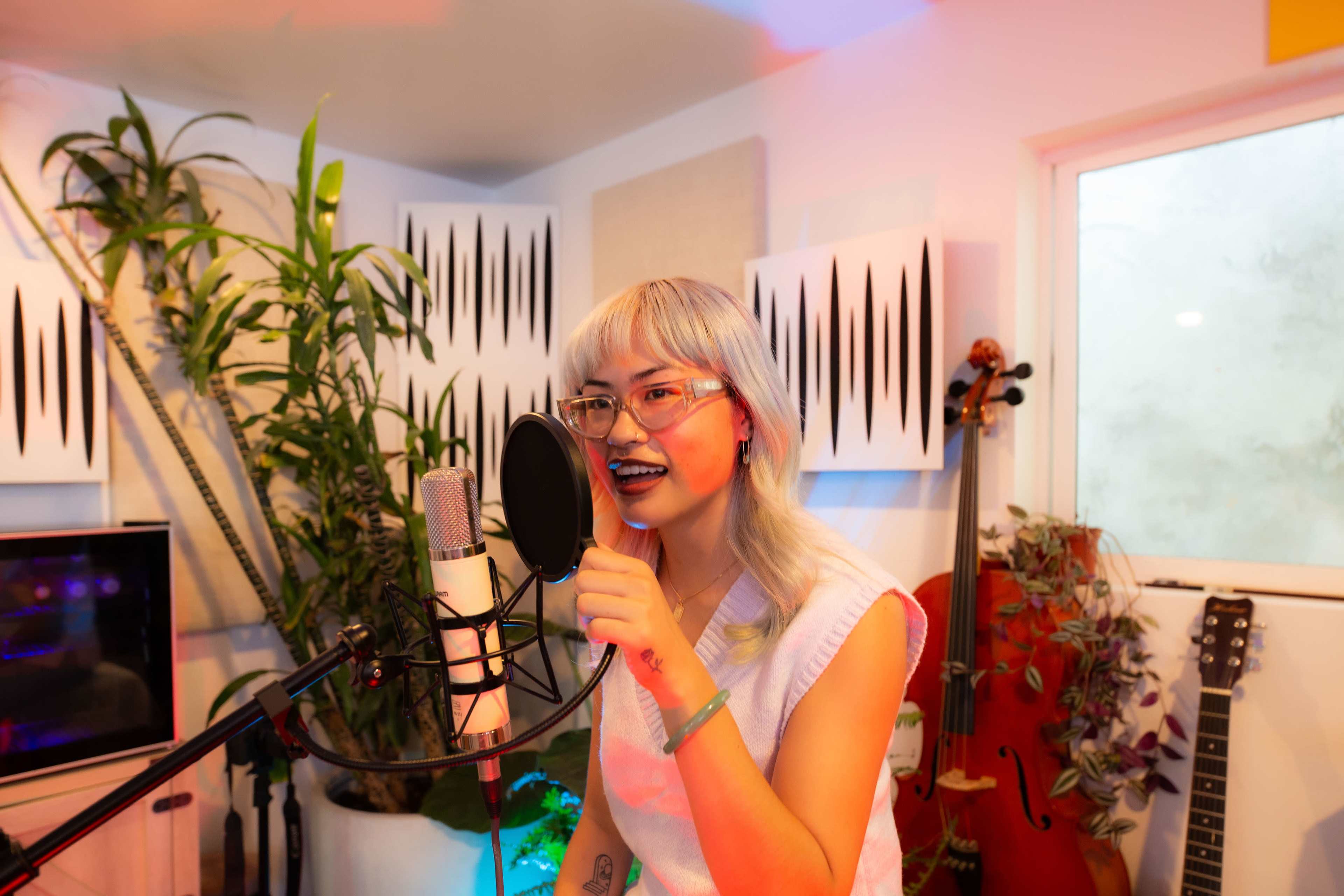 A young woman with light-colored hair is singing into a microphone in a brightly lit recording studio surrounded by plants and musical instruments.
