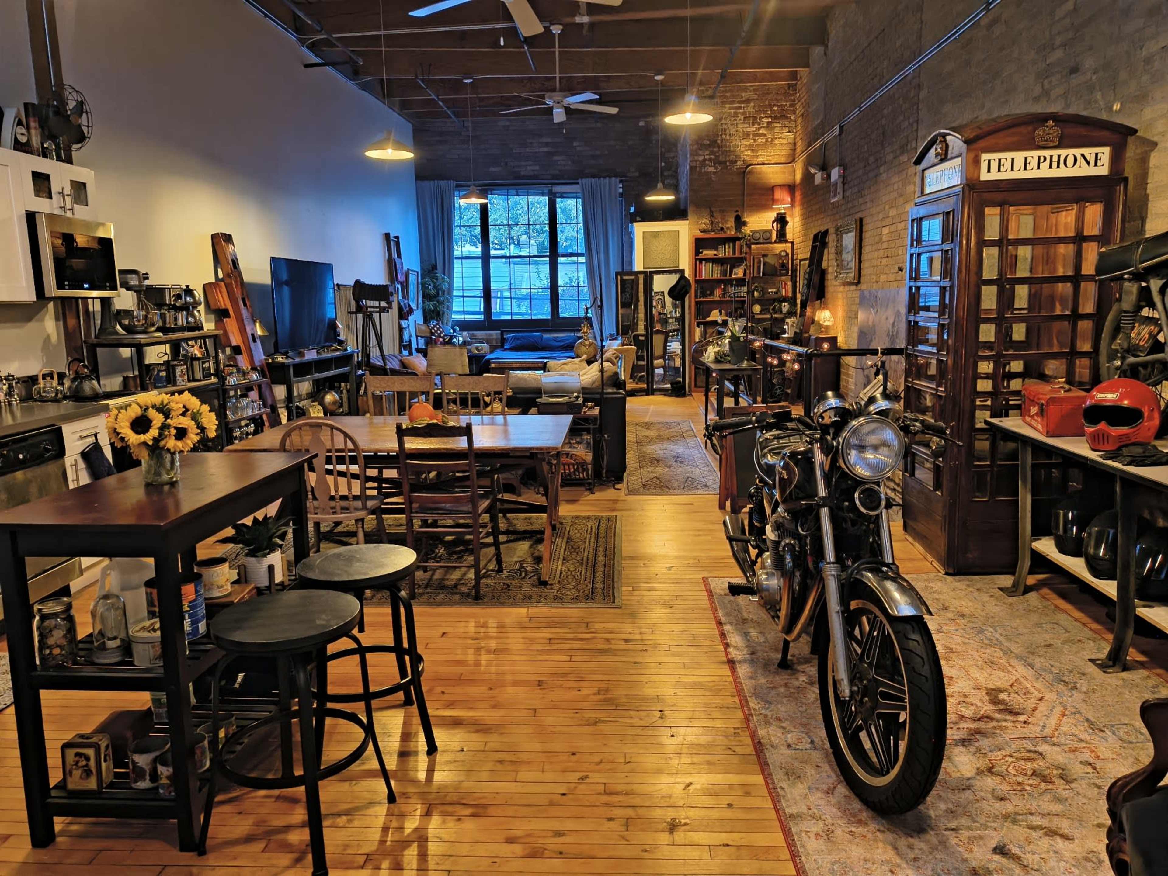 The image shows a spacious loft with a motorcycle on a rug, surrounded by wooden furniture, shelves, and a cozy seating area near a large window.