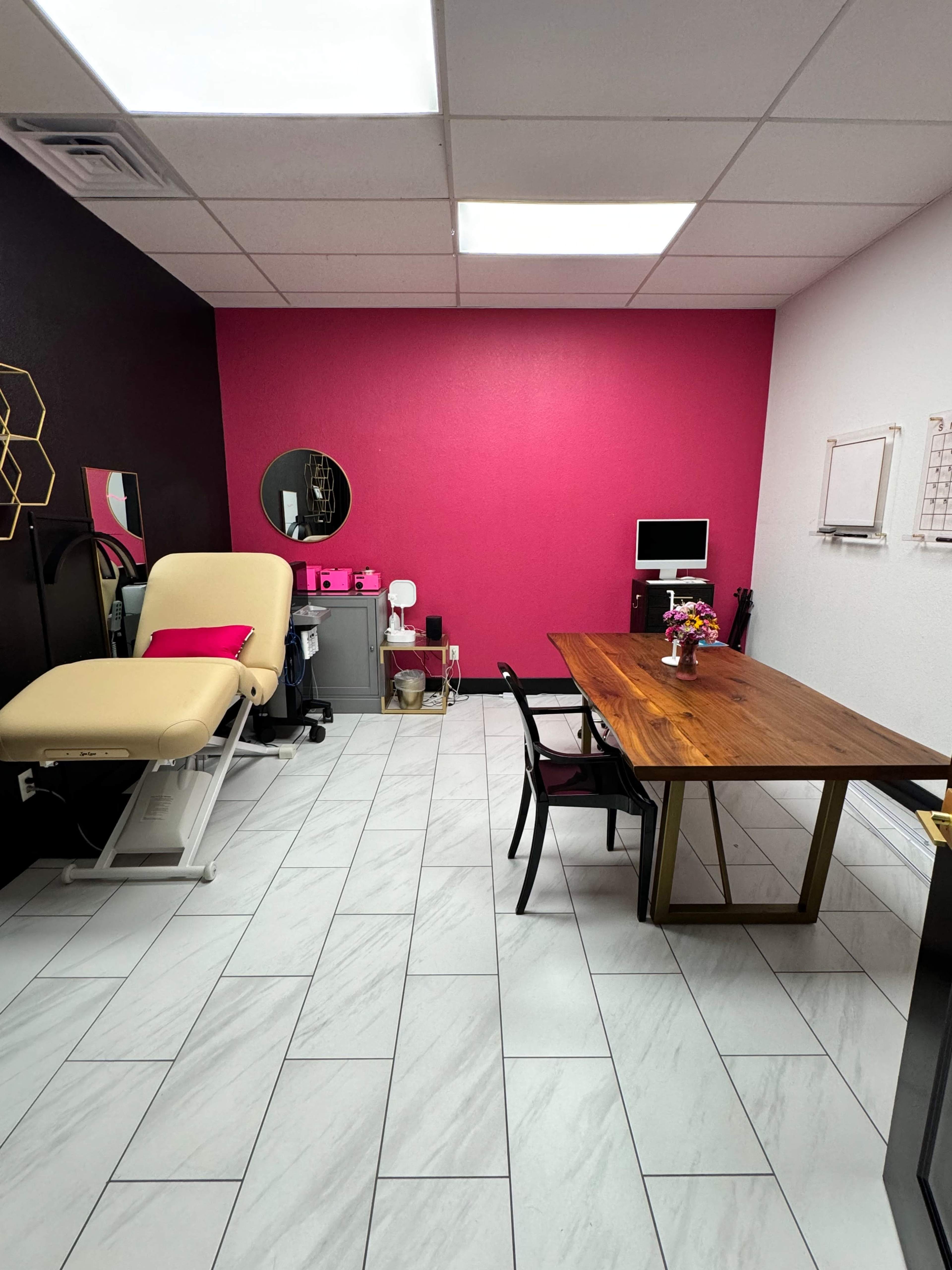 The image shows a medical or treatment room featuring a cream-colored examination chair, a wooden table with chairs, and a pink accent wall.